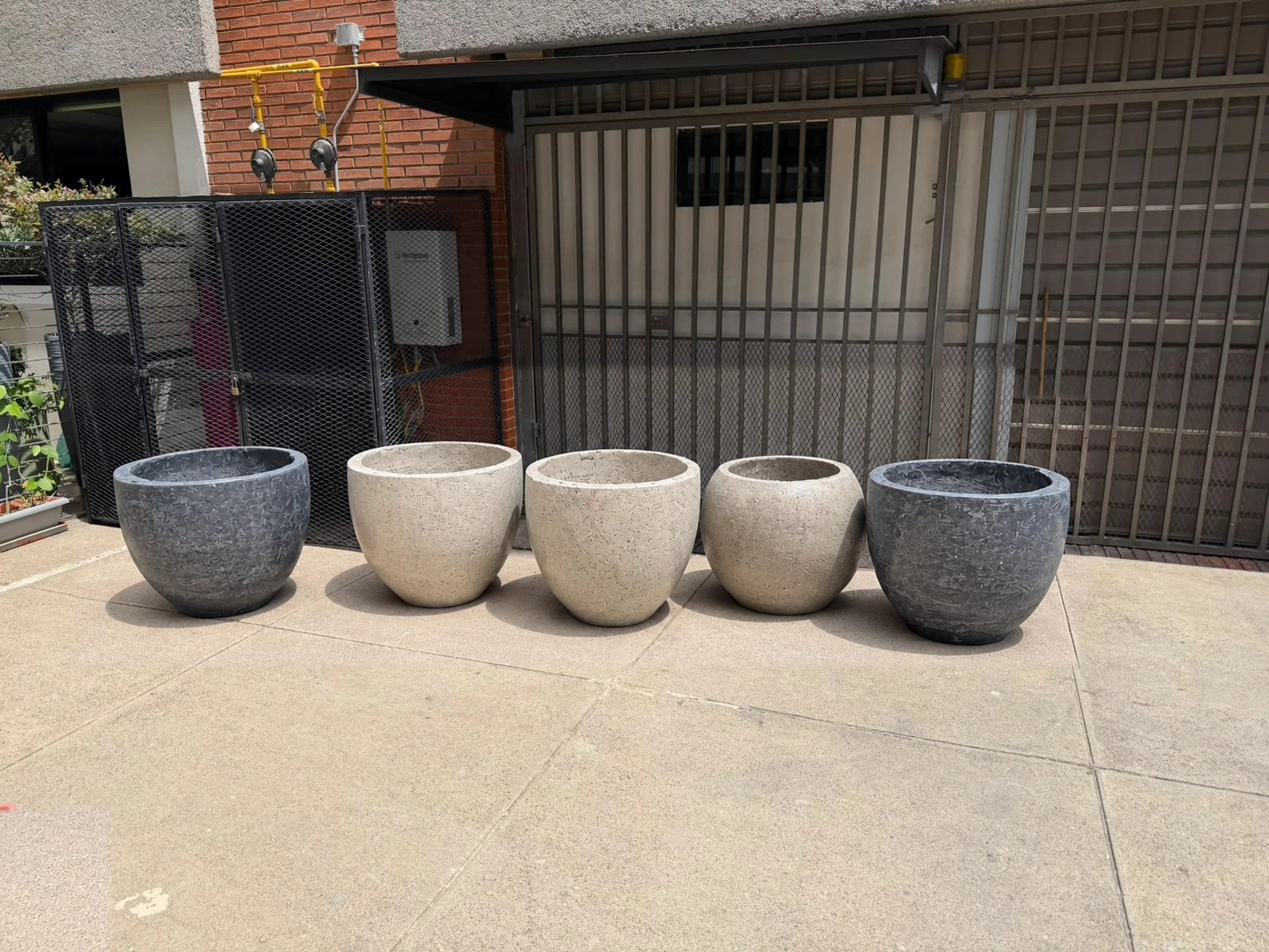 Cinco maceteros redondos de concreto, grises y claros, alineados sobre piso de cemento frente a una reja