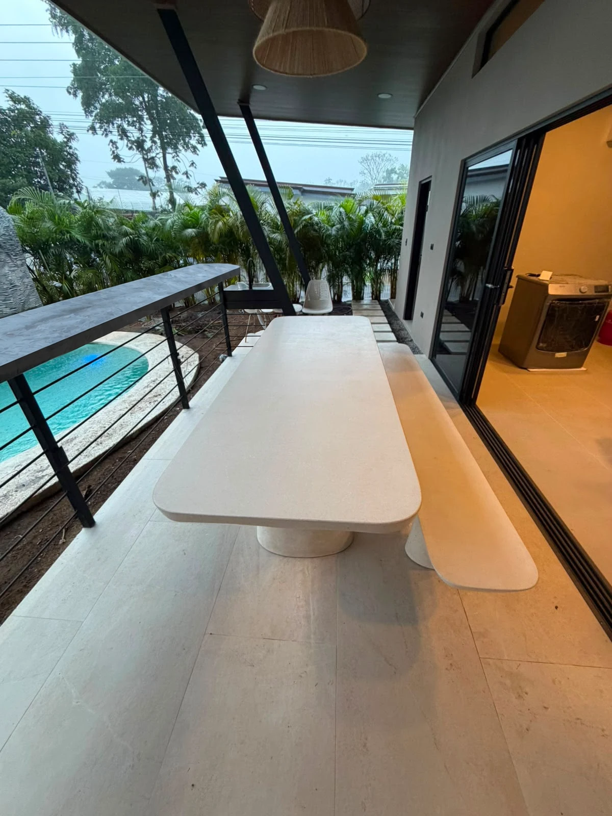 Mesa de comedor de concreto color crema con borde redondeado y banca a juego, en terraza techada frente a la piscina