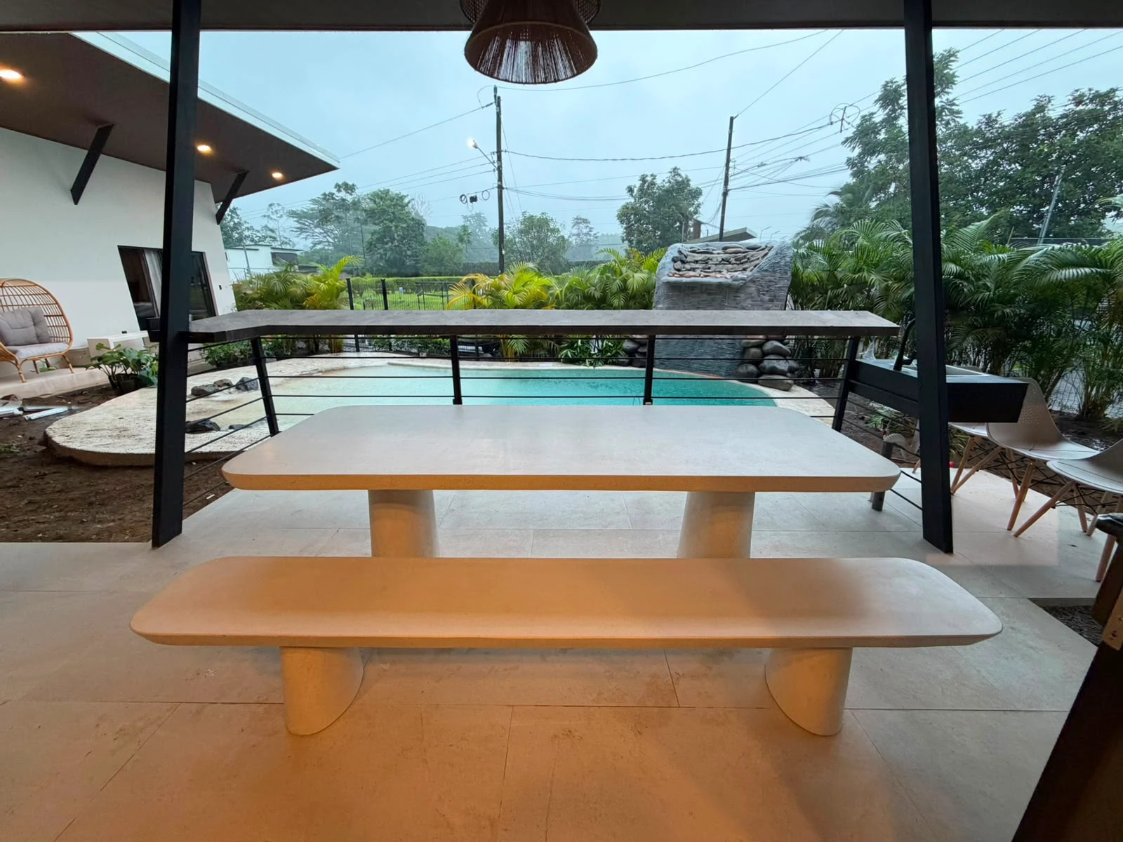Mesa de comedor de concreto color arena con banca larga a juego, bajo techo en terraza abierta con vista al jardín y la piscina