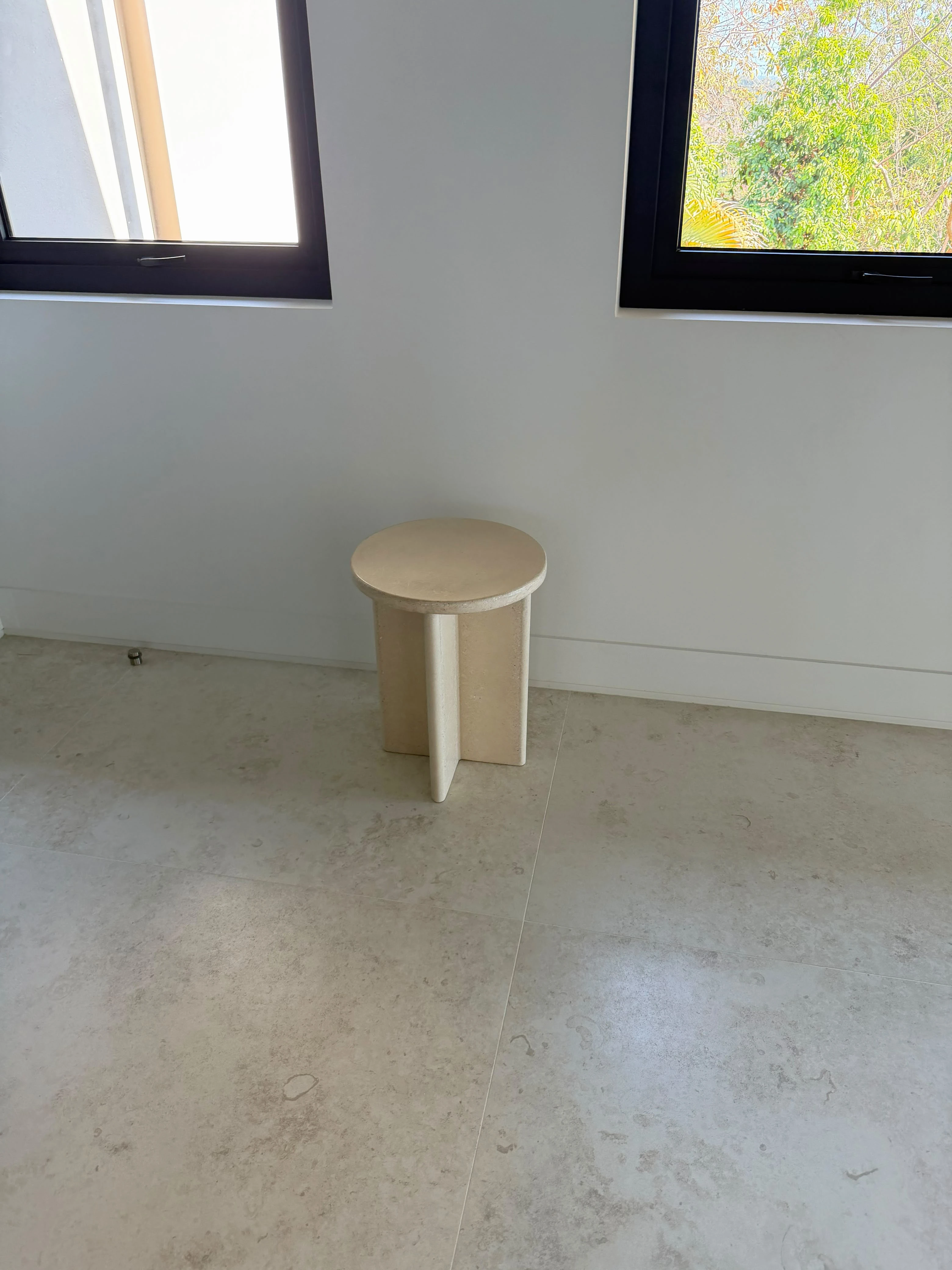 Banco o mesa auxiliar redonda de concreto color crema con tres patas en forma de aleta, sobre piso pulido en habitación luminosa