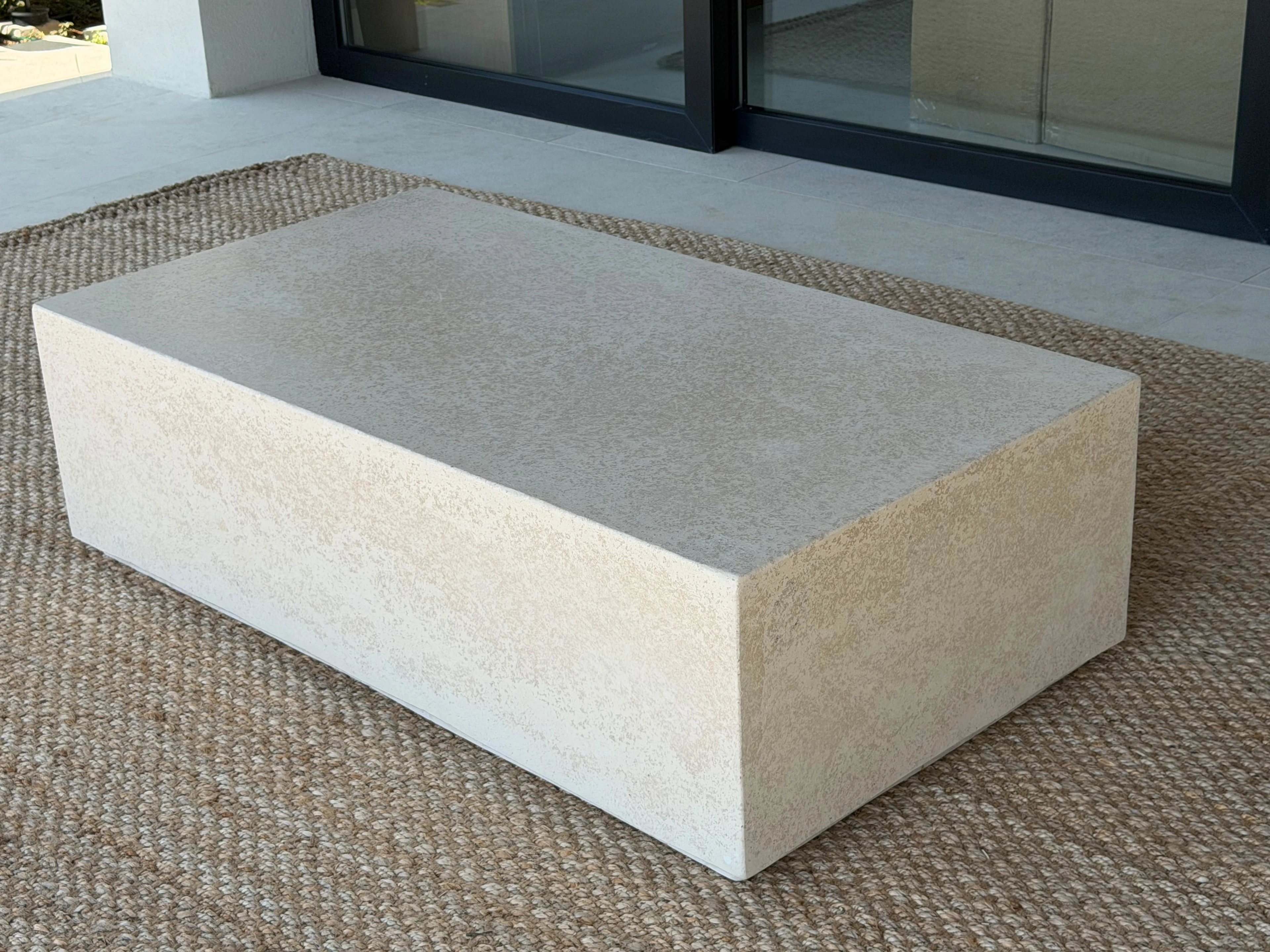 Mesa de centro rectangular de concreto color crema con tapa más oscura y laterales granulados, sobre alfombra de fibra natural