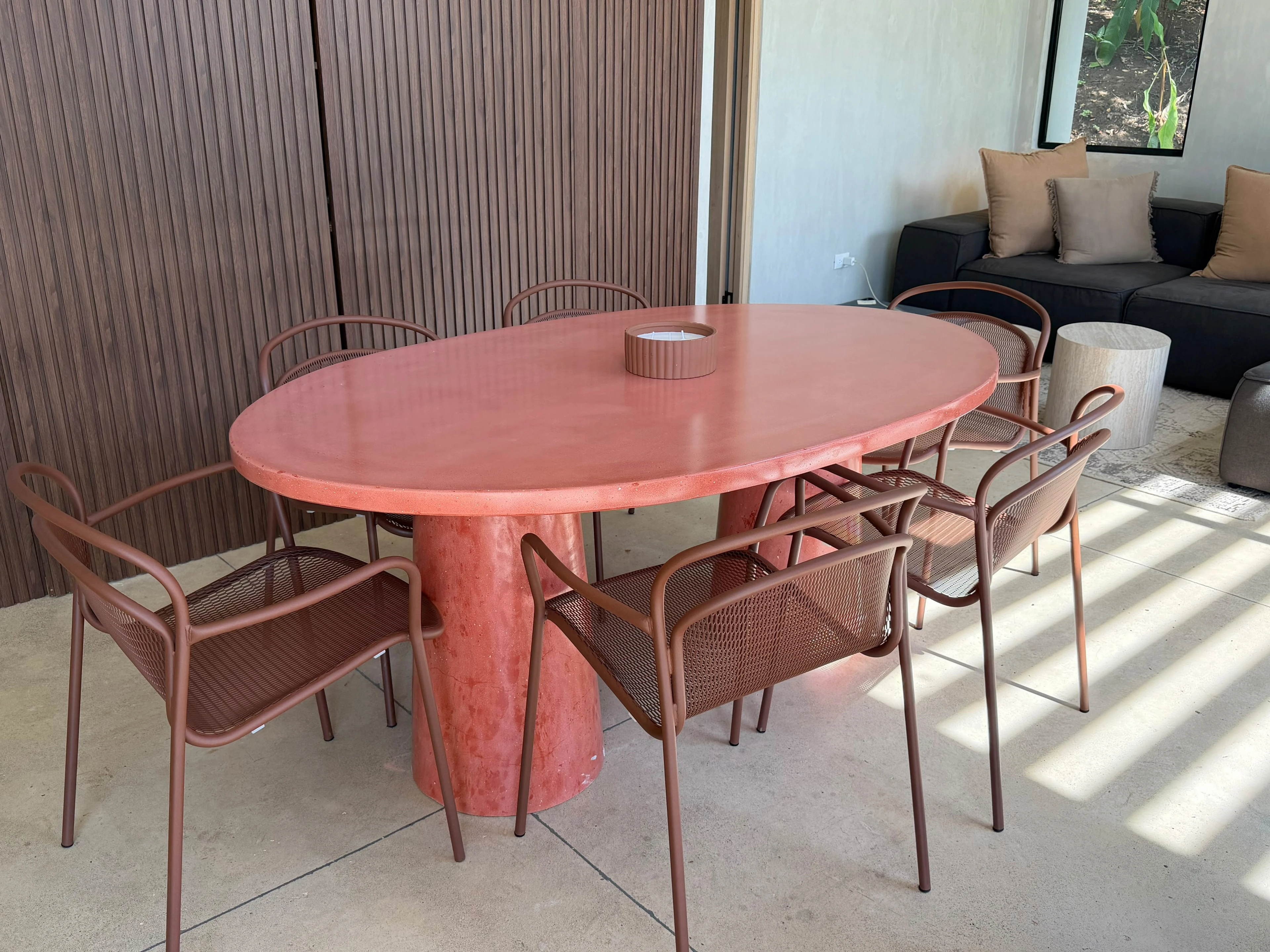 Mesa de comedor ovalada de concreto terracota con pedestal central, rodeada de sillas metálicas en sala con paneles de madera