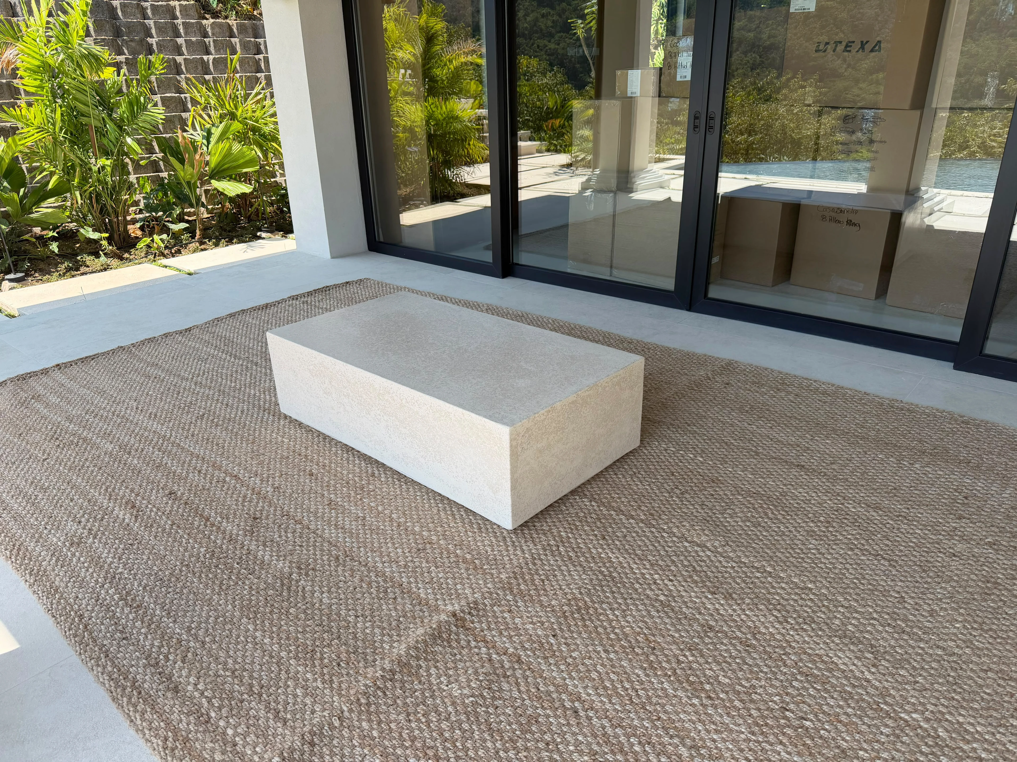 Mesa de centro baja y rectangular de concreto color crema sobre alfombra de fibra natural, frente a ventanales