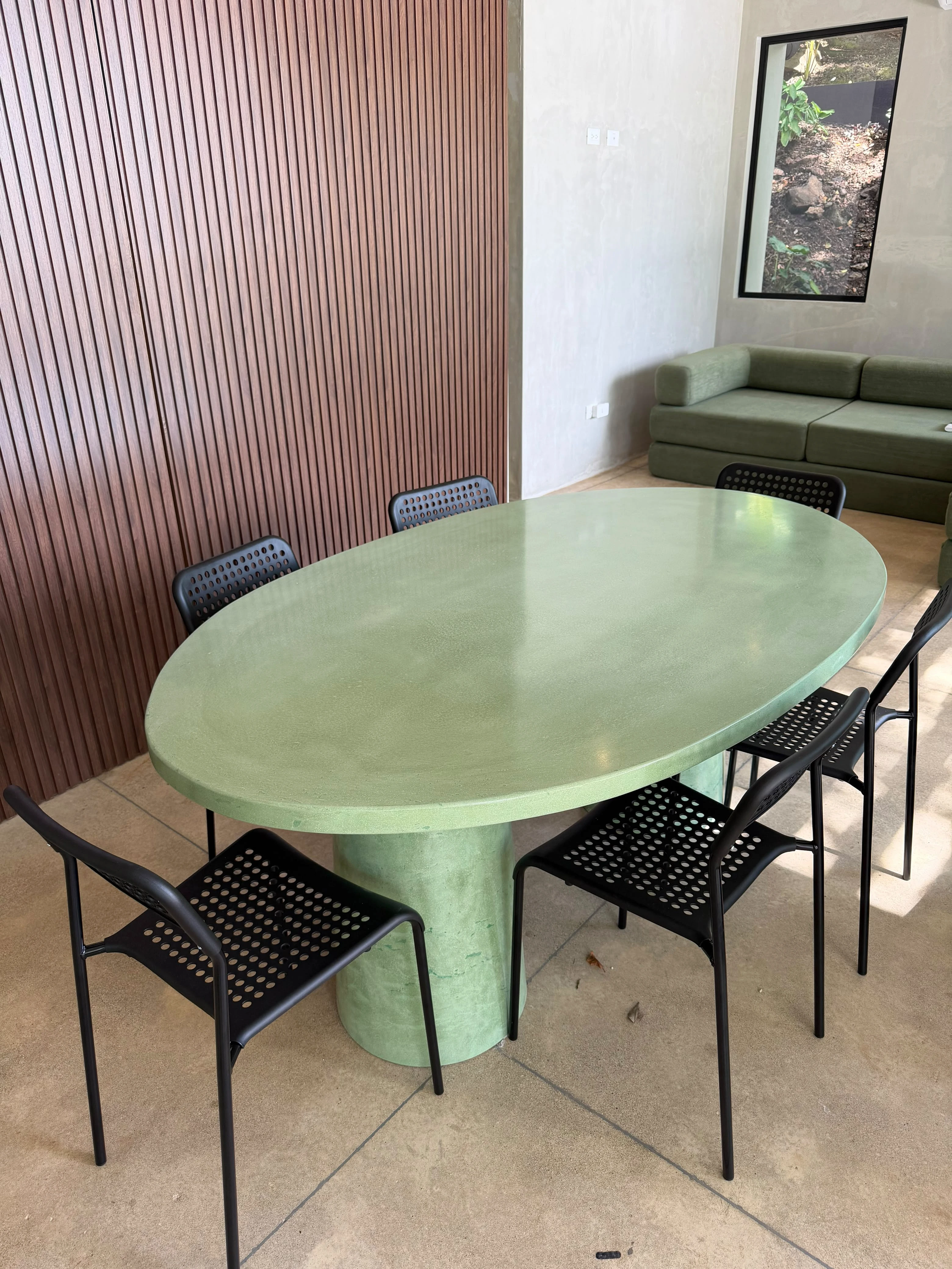 Mesa de comedor ovalada de concreto verde pastel con pedestal central y sillas negras, en sala con paneles de madera
