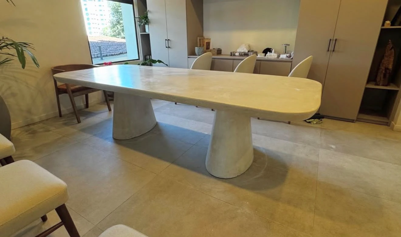 Mesa de comedor larga de concreto color crema con bordes redondeados y dos patas cónicas, en comedor interior