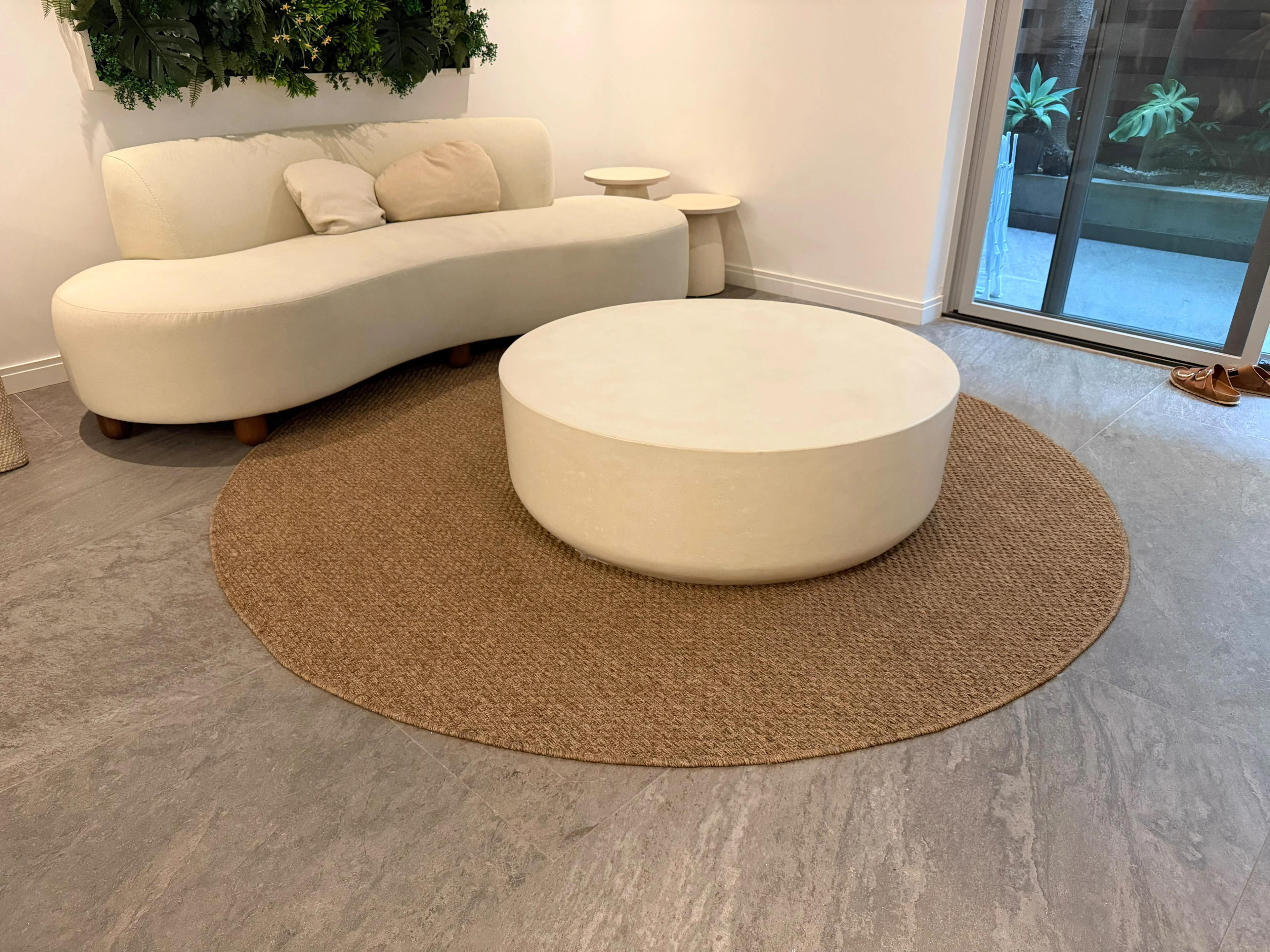 Mesa de centro redonda y baja de concreto blanco crema sobre alfombra circular de fibra, junto a sofá curvo en sala