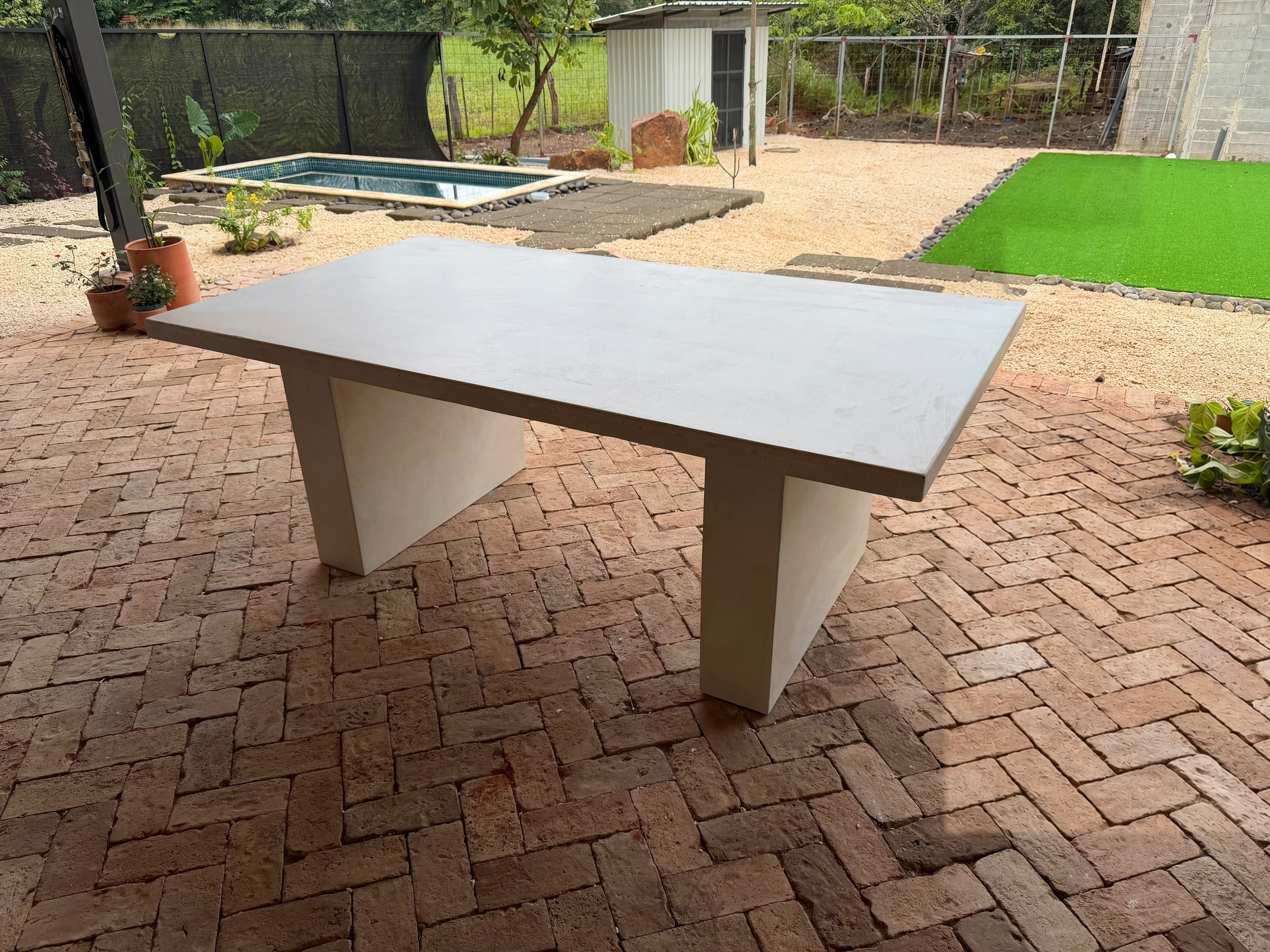 Mesa rectangular de concreto gris con patas laterales inclinadas, sobre piso de ladrillo en patio con césped y piscina