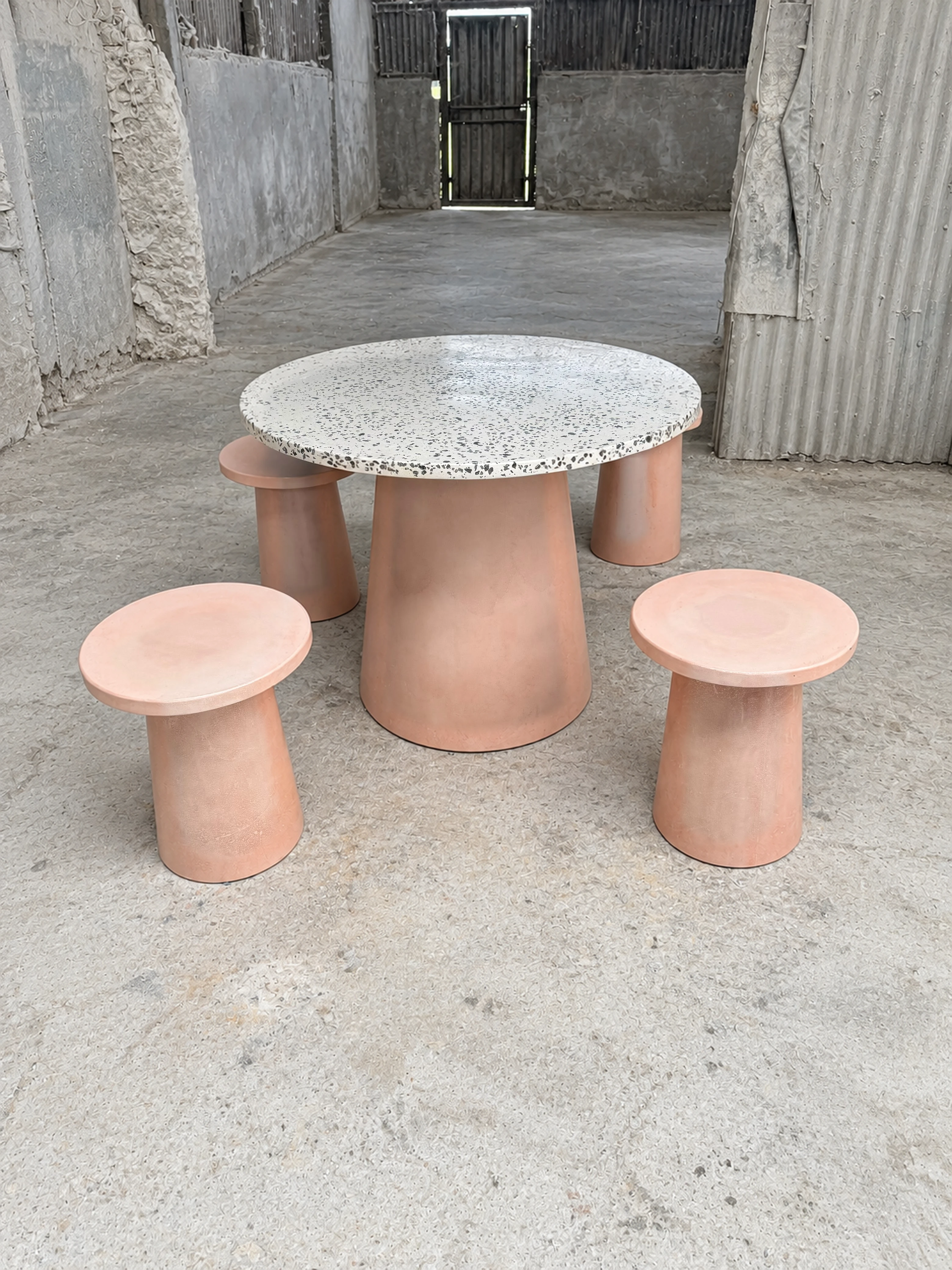 Mesa redonda de terrazo blanco con agregados oscuros y tres banquitos de concreto rosado en forma de hongo, en bodega con piso pulido