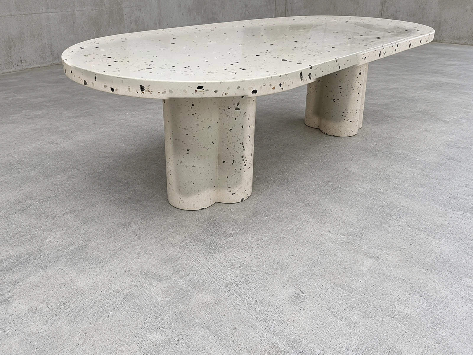 Mesa baja ovalada de terrazo blanco con agregados oscuros y patas en forma de trébol, sobre piso de cemento pulido