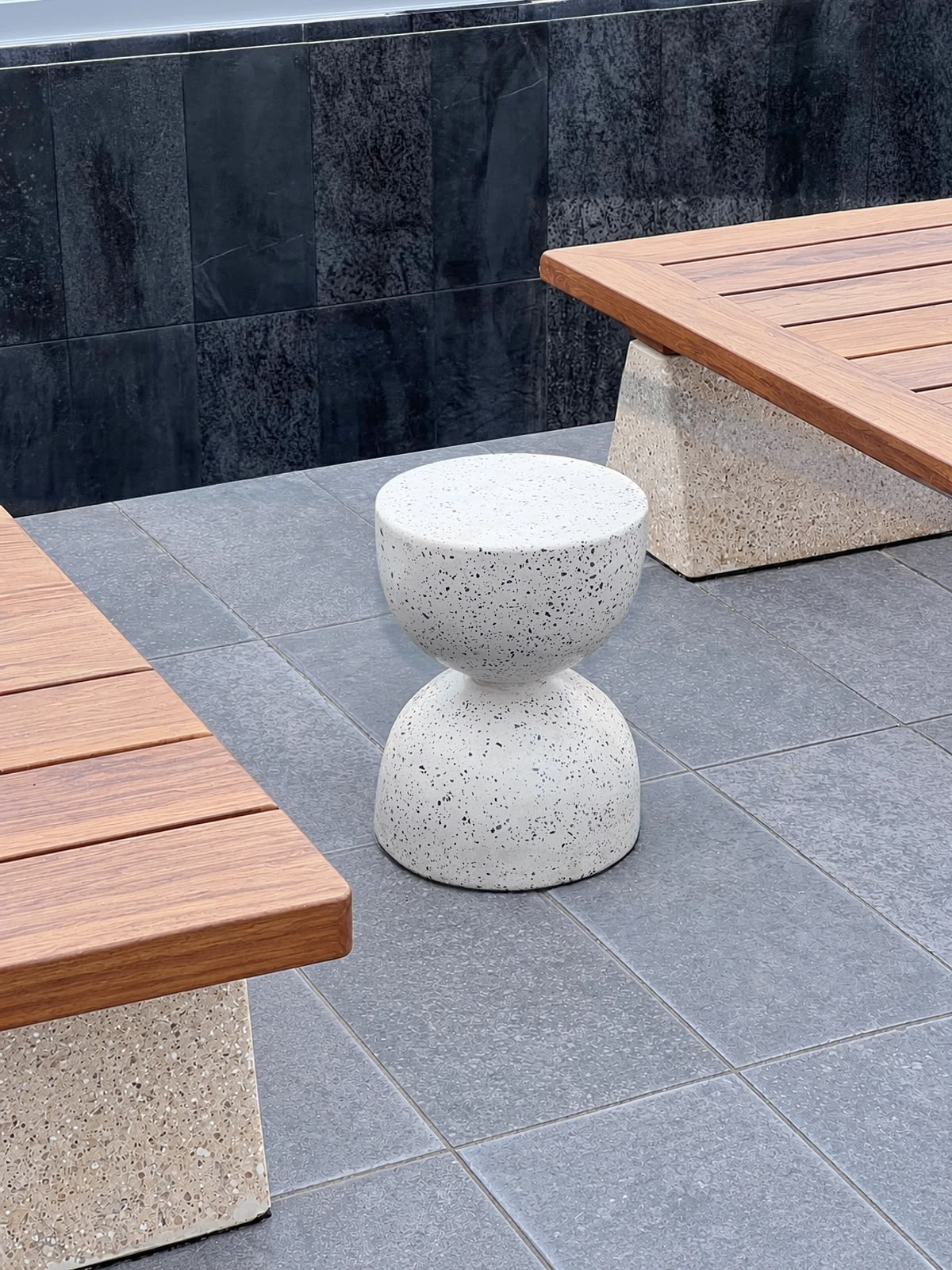 Banquito de terrazo blanco con agregados negros en forma de reloj de arena, junto a bancas de madera con base de concreto