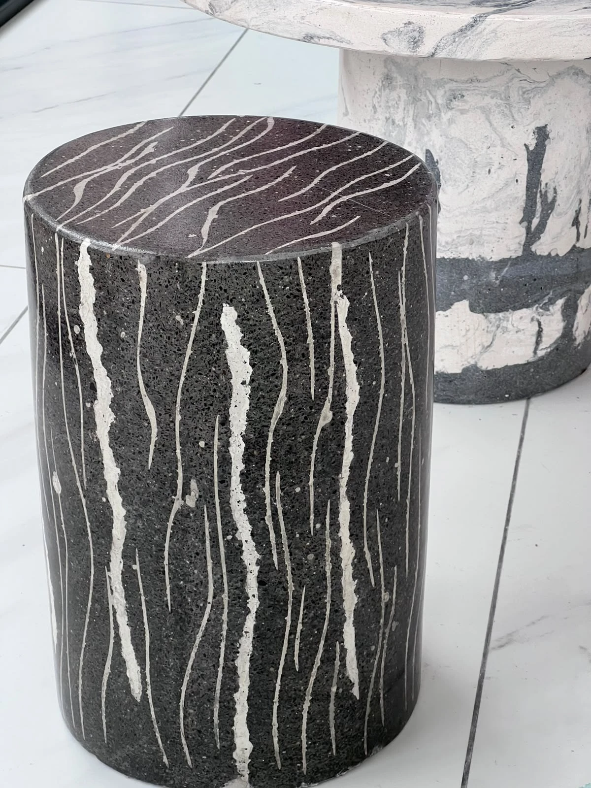 Banco cilíndrico de terrazo negro con vetas blancas onduladas a modo de rayas, sobre piso de porcelanato blanco
