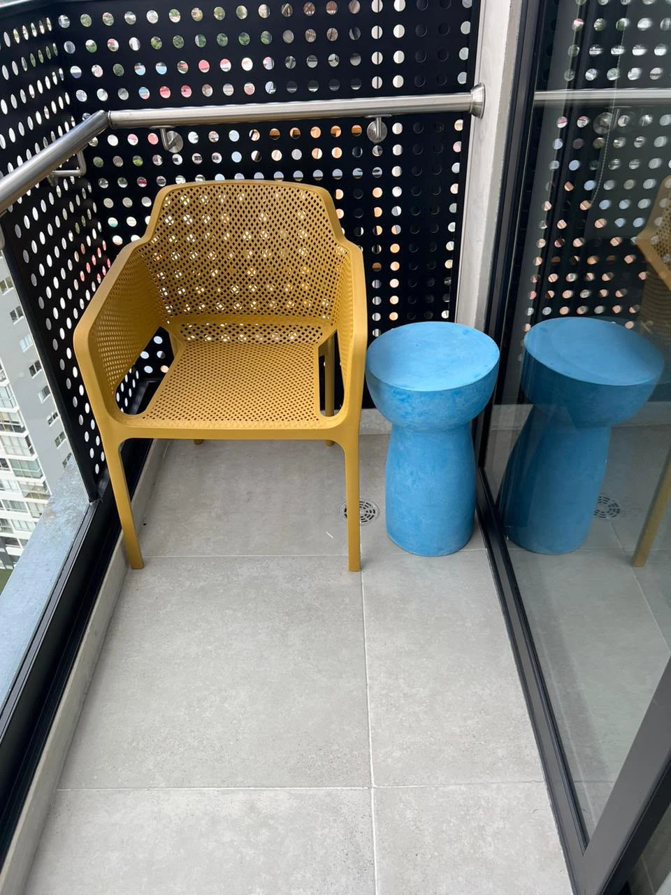 Dos banquitos de concreto azul con forma de reloj de arena junto a una silla amarilla, en balcón con barandal perforado