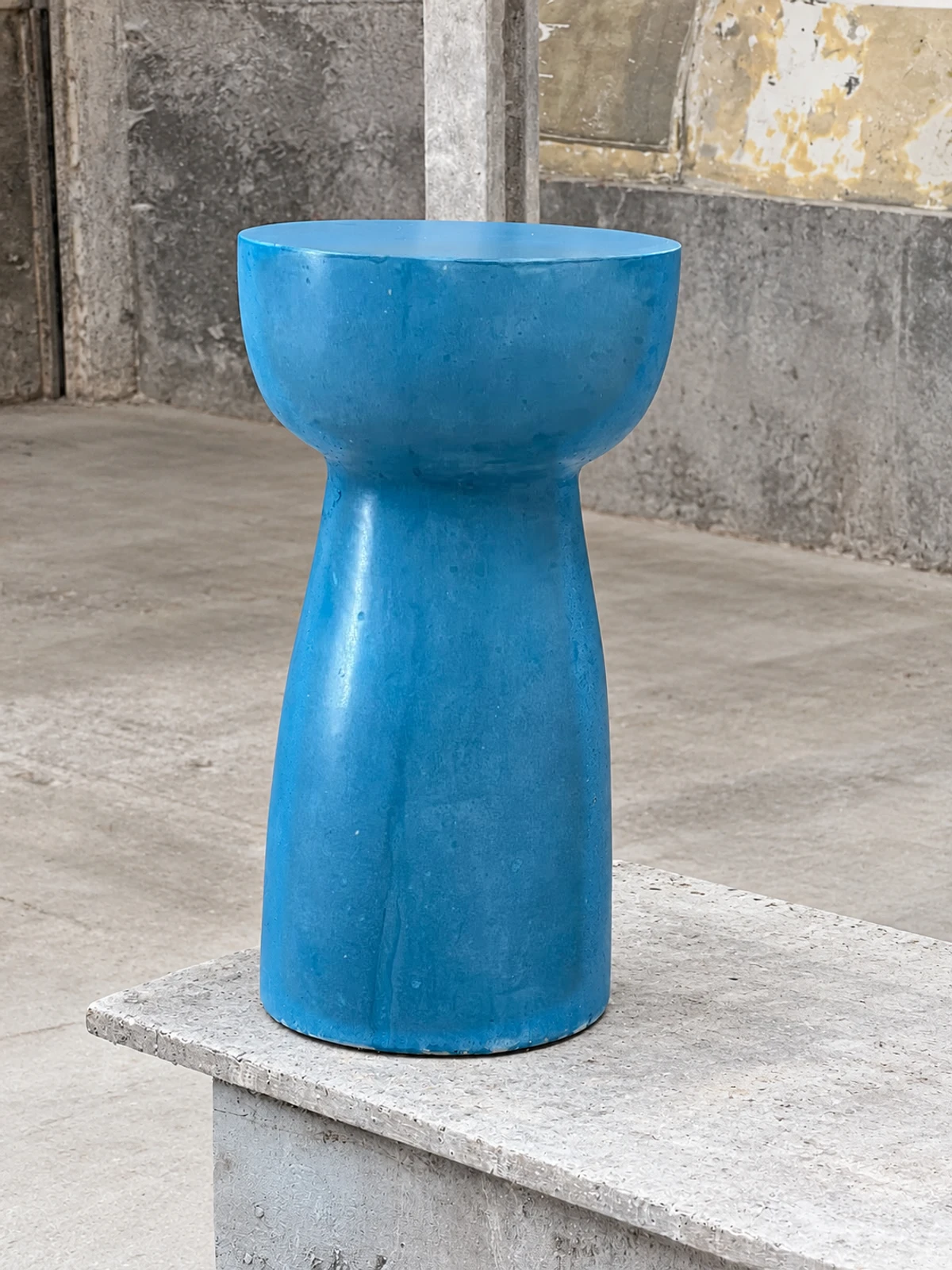 Mesa auxiliar alta de concreto azul intenso con tapa redonda y pie estrecho, sobre una base de piedra en espacio industrial