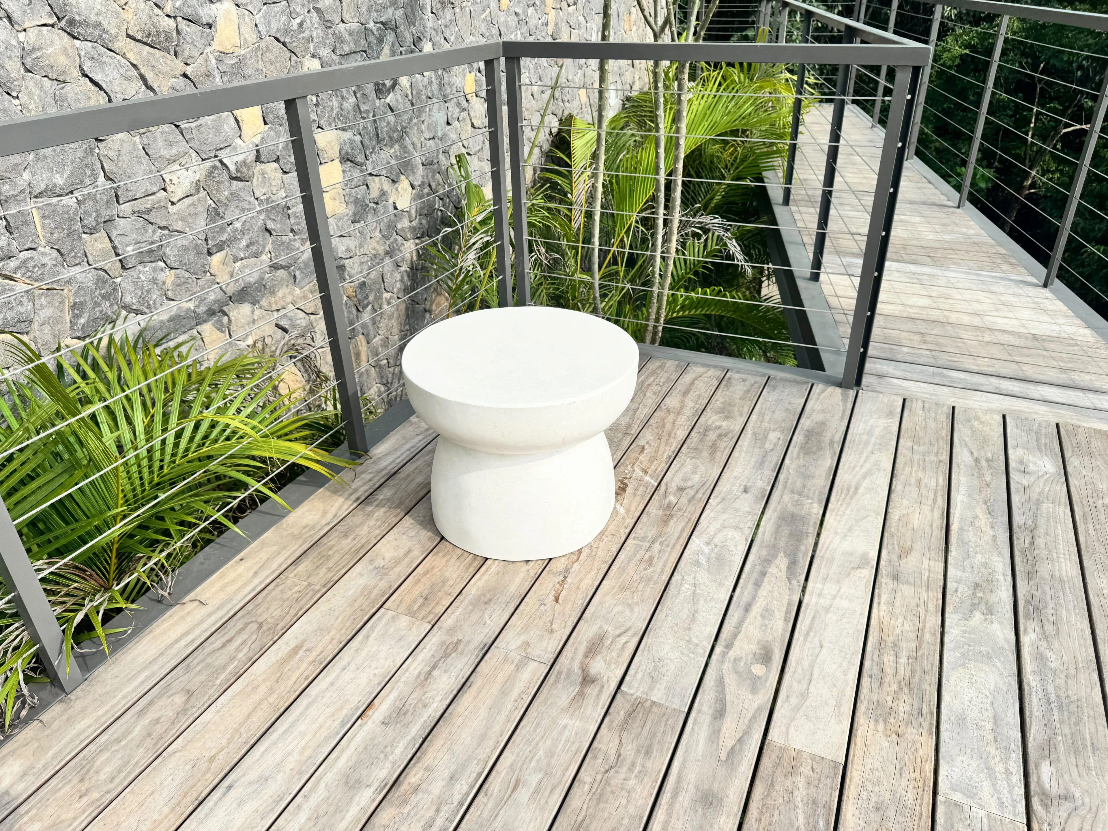 Mesa auxiliar baja de concreto blanco con forma de hongo, sobre deck de madera en terraza con baranda de cables y palmas
