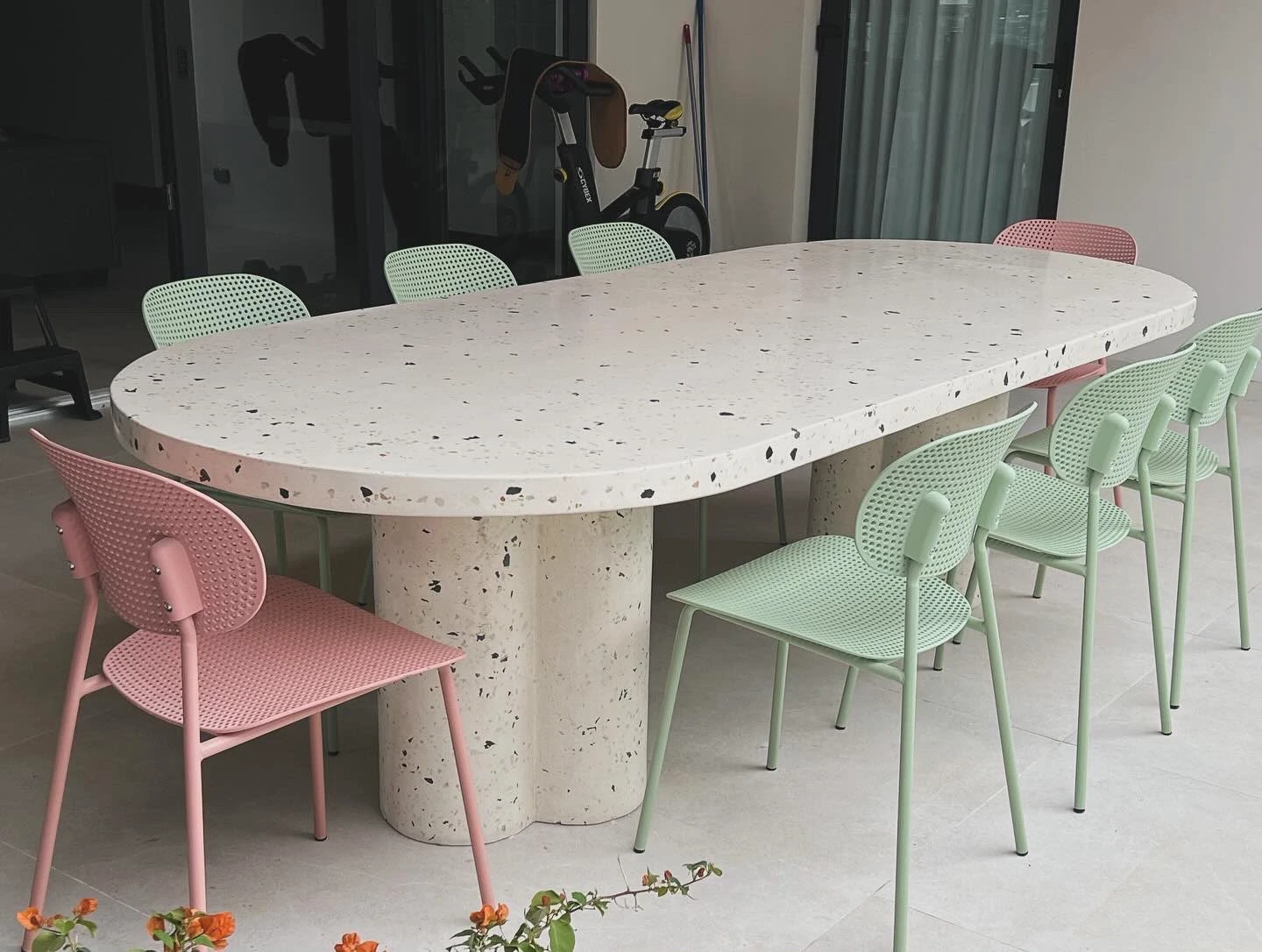 Mesa de comedor ovalada de terrazo blanco con agregados oscuros, pata central en forma de trébol y sillas rosadas y verdes