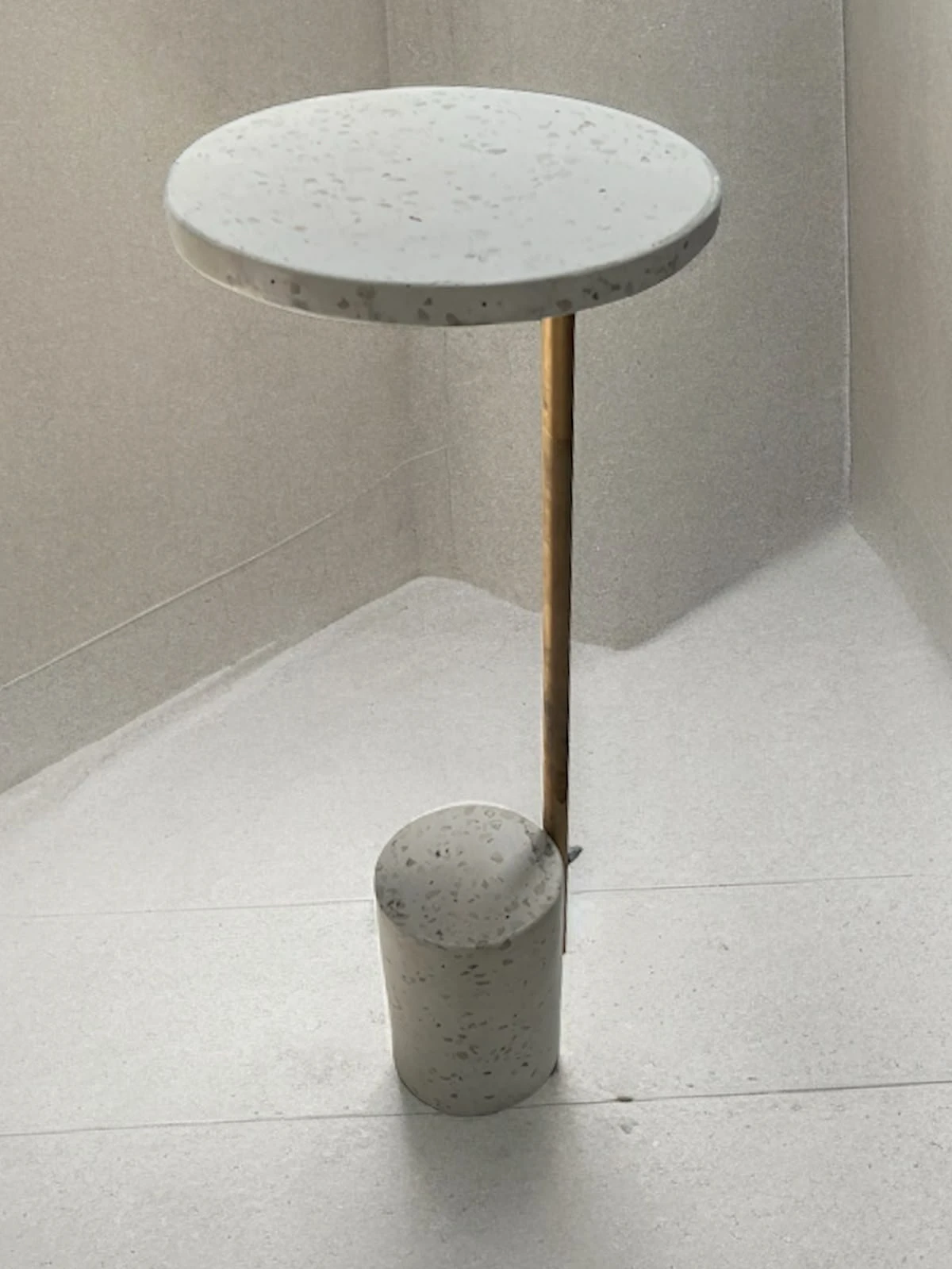 Mesa auxiliar de terrazo blanco con tapa circular delgada, vástago de latón y base cilíndrica, sobre piso claro