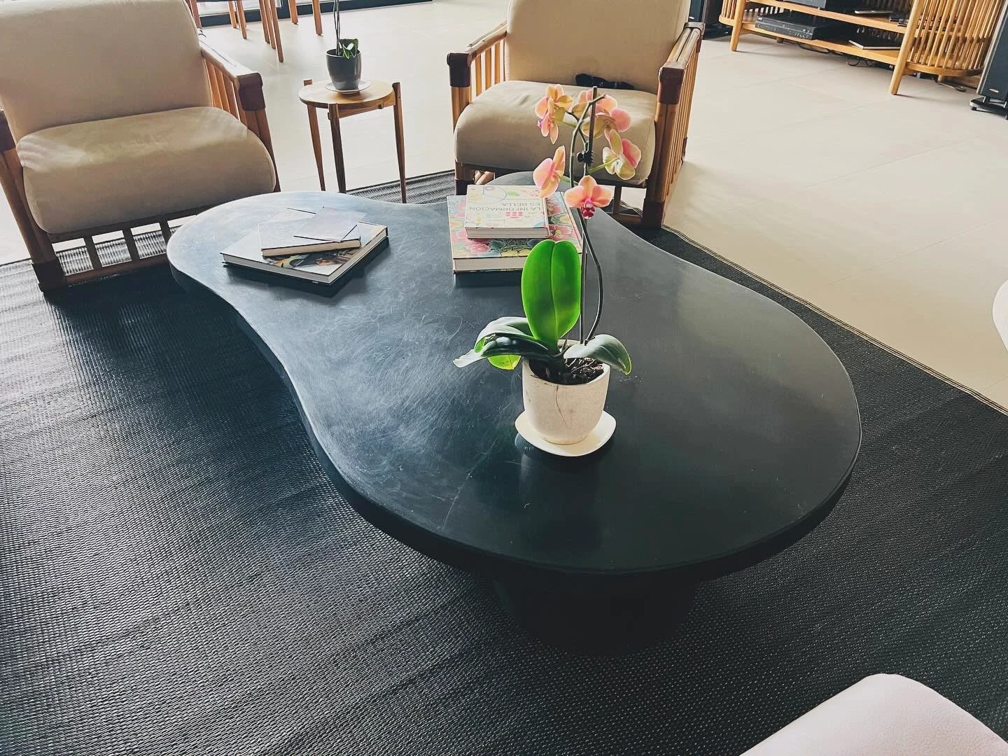 Mesa de centro de concreto negro con forma orgánica de frijol, con libros y una orquídea, en sala con sillones y alfombra oscura