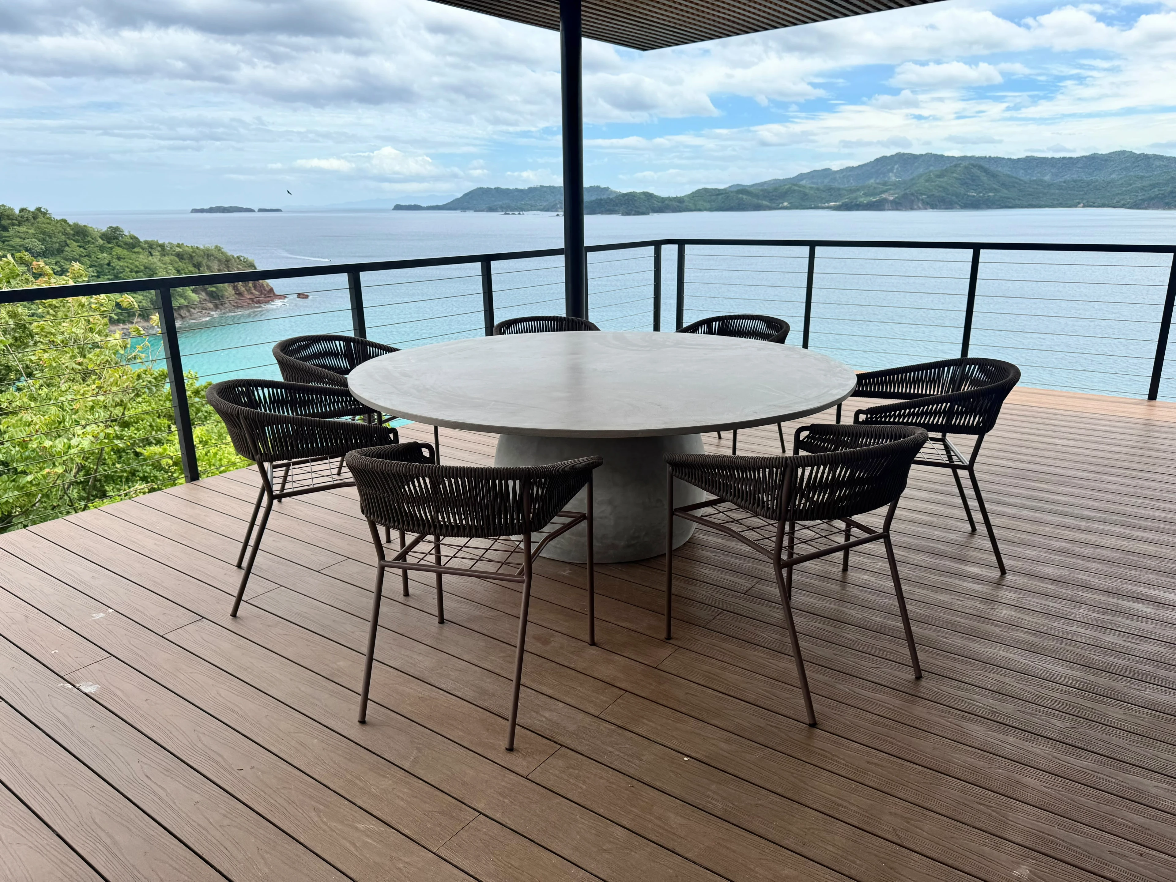 Mesa de comedor redonda de concreto claro con pata central y sillas de cuerda negras, en terraza de deck con vista al mar
