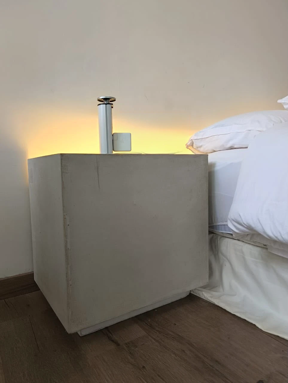 Mesa de noche cúbica de concreto gris liso junto a una cama, con luz cálida en la pared y piso de madera