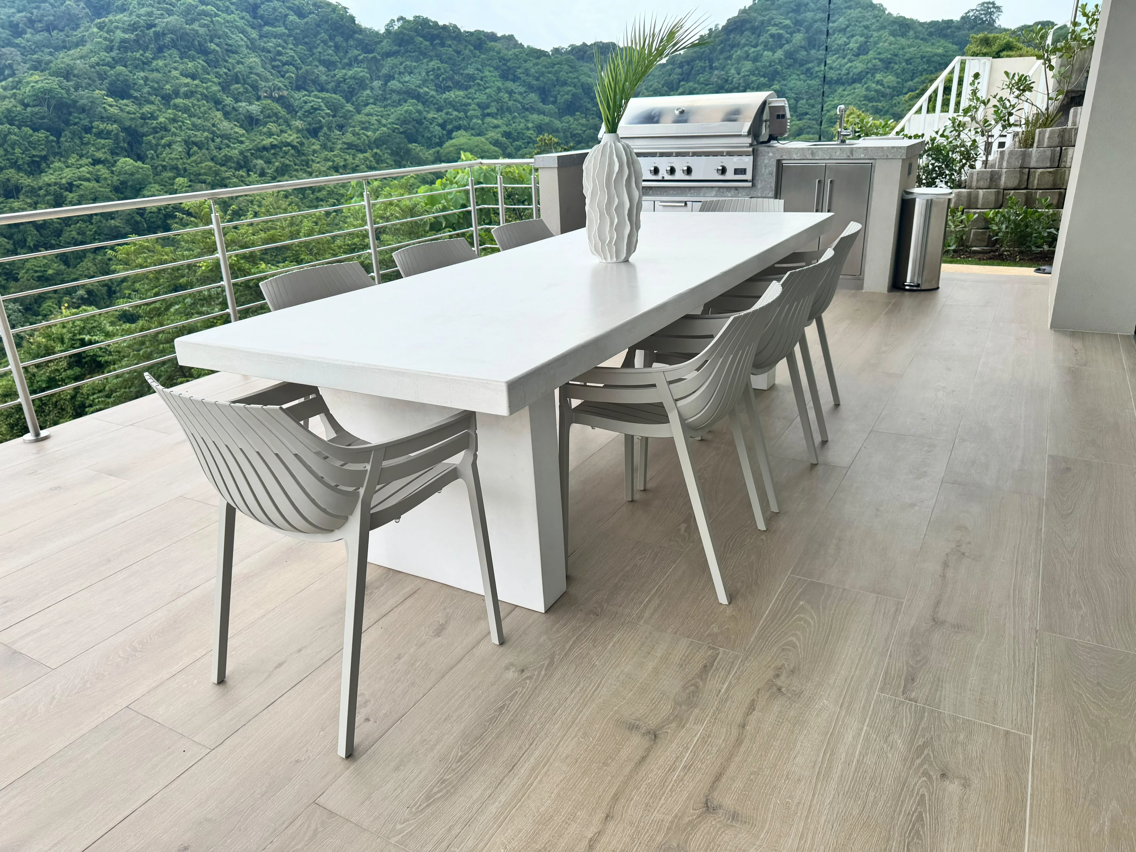 Mesa de comedor larga de concreto blanco con patas laterales, sillas grises y barbacoa, en terraza con vista a la montaña