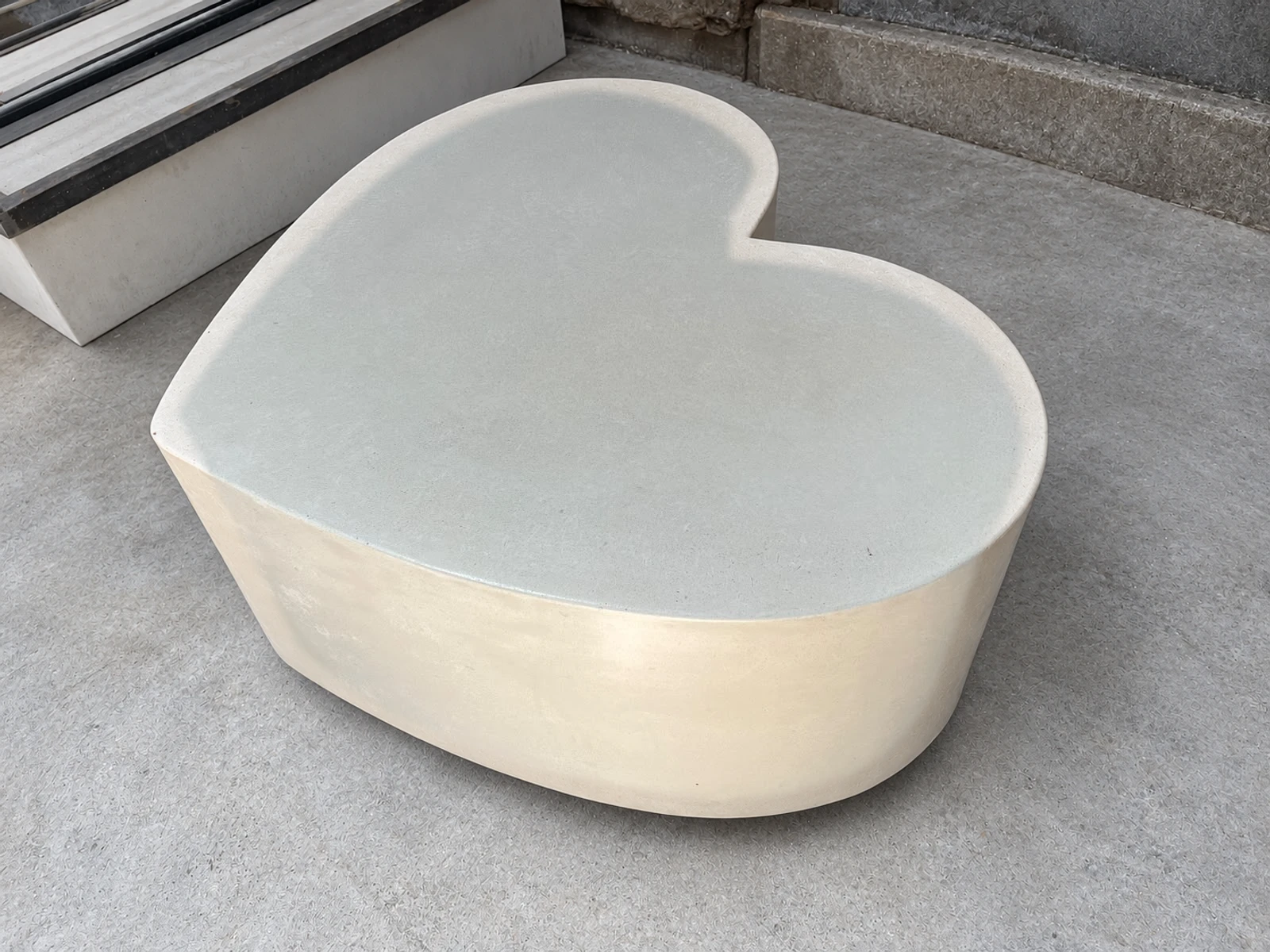 Mesa baja con forma de corazón de concreto claro con tapa lisa gris azulada y laterales crema, sobre piso de cemento
