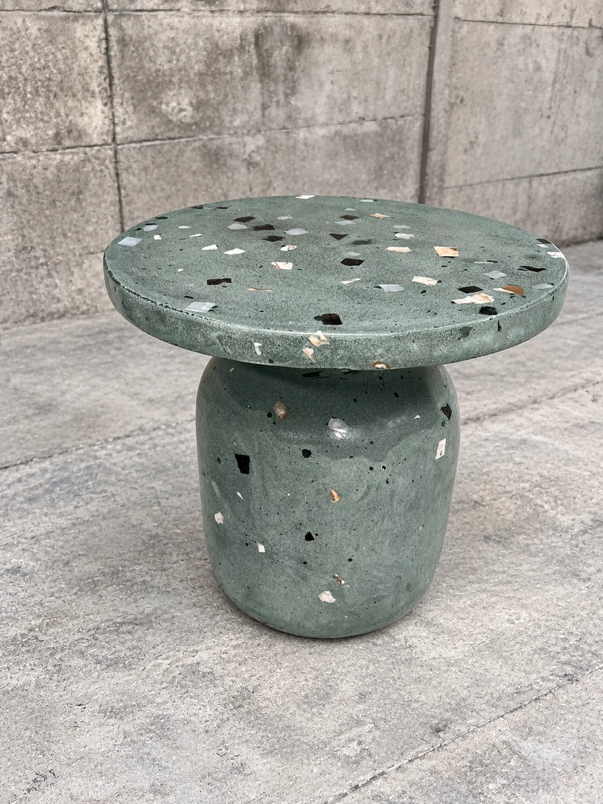 Mesa auxiliar de terrazo verde con agregados de colores, tapa redonda y pie abombado, sobre piso de cemento pulido