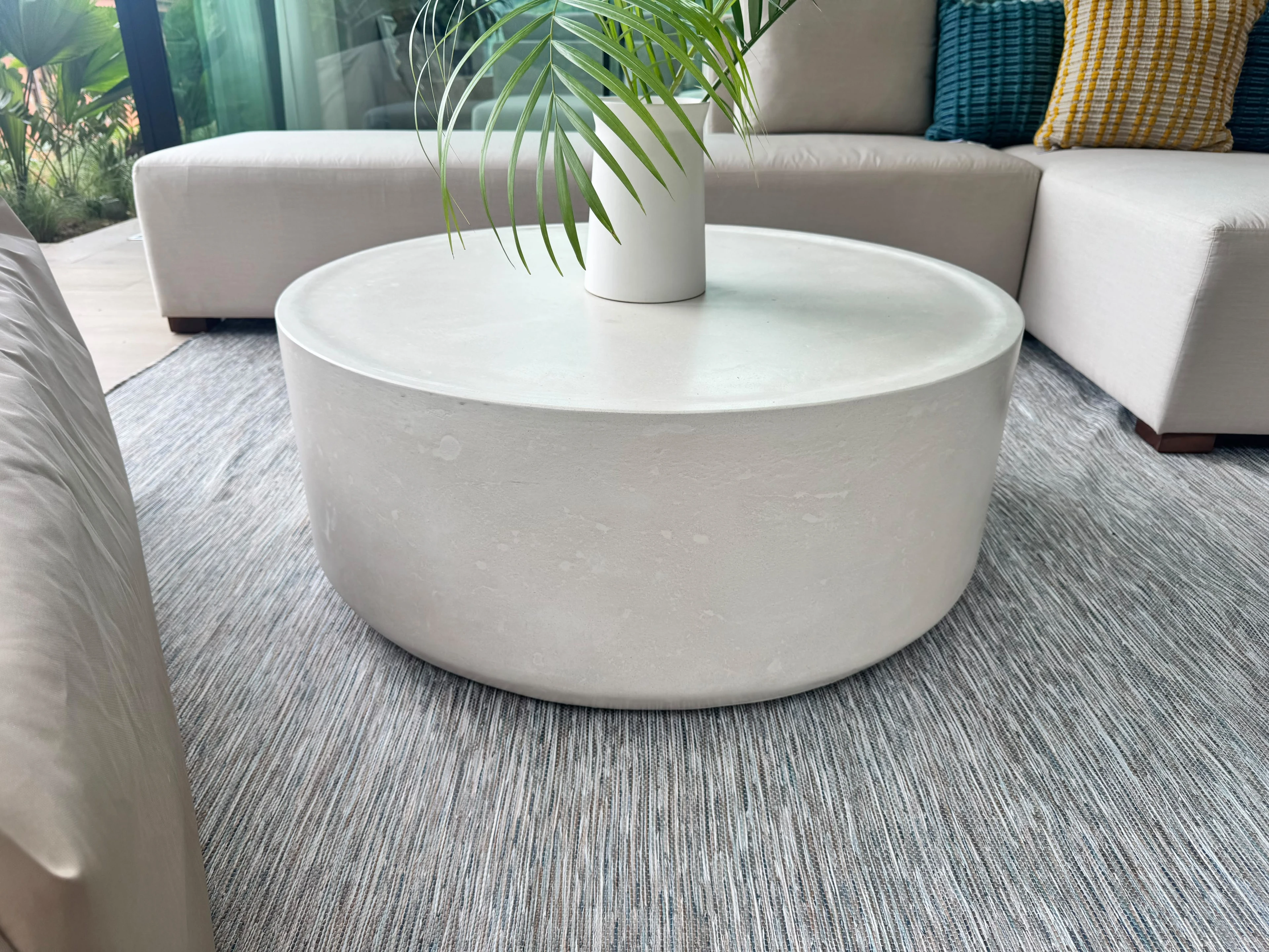 Mesa de centro redonda de concreto blanco con borde recto y una planta encima, sobre alfombra gris frente a un sofá