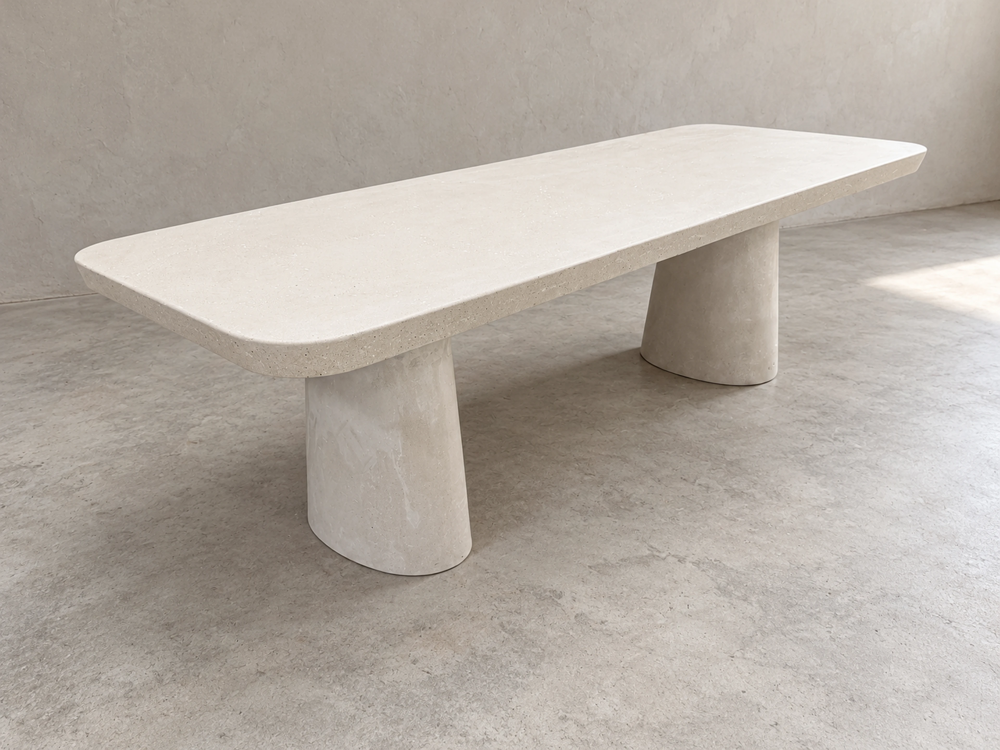 Mesa de comedor larga de concreto blanco con dos patas cónicas y esquinas redondeadas, sobre piso liso de tono claro