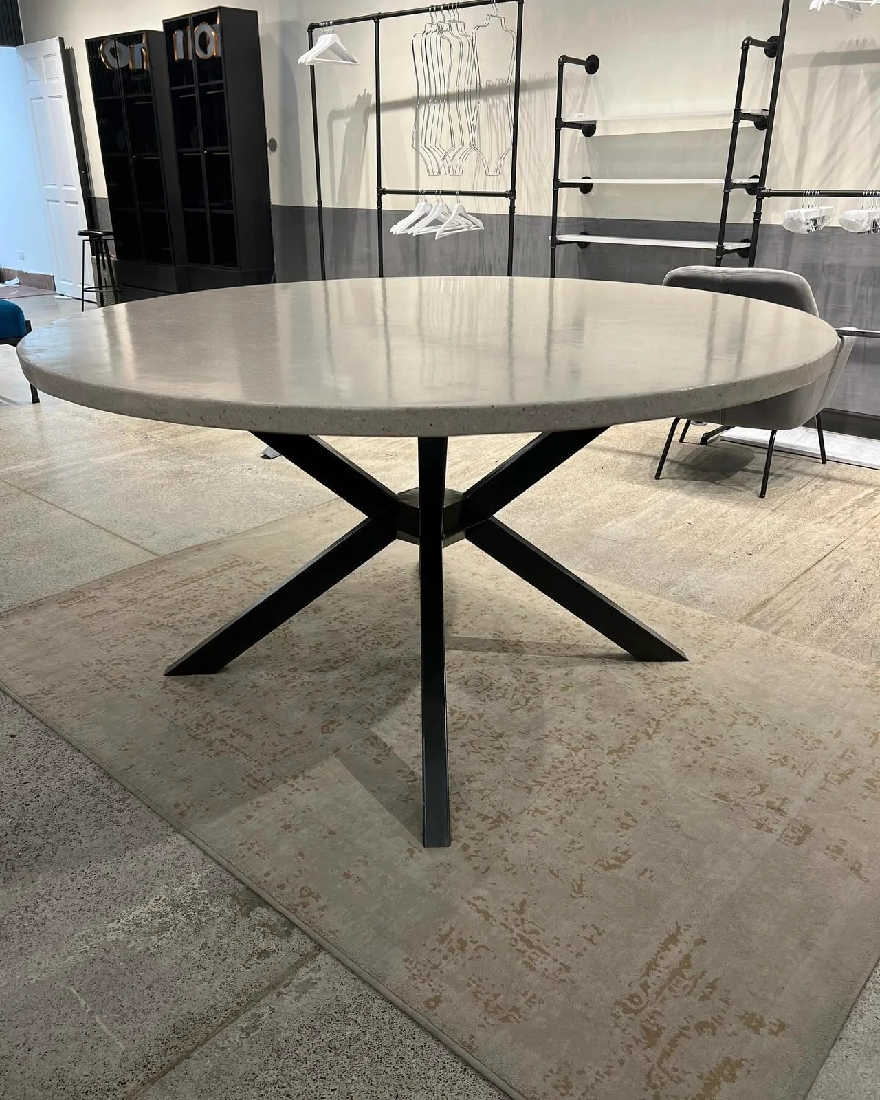 Mesa de comedor redonda con tapa de concreto gris claro y base de metal negro cruzada, sobre alfombra en interior