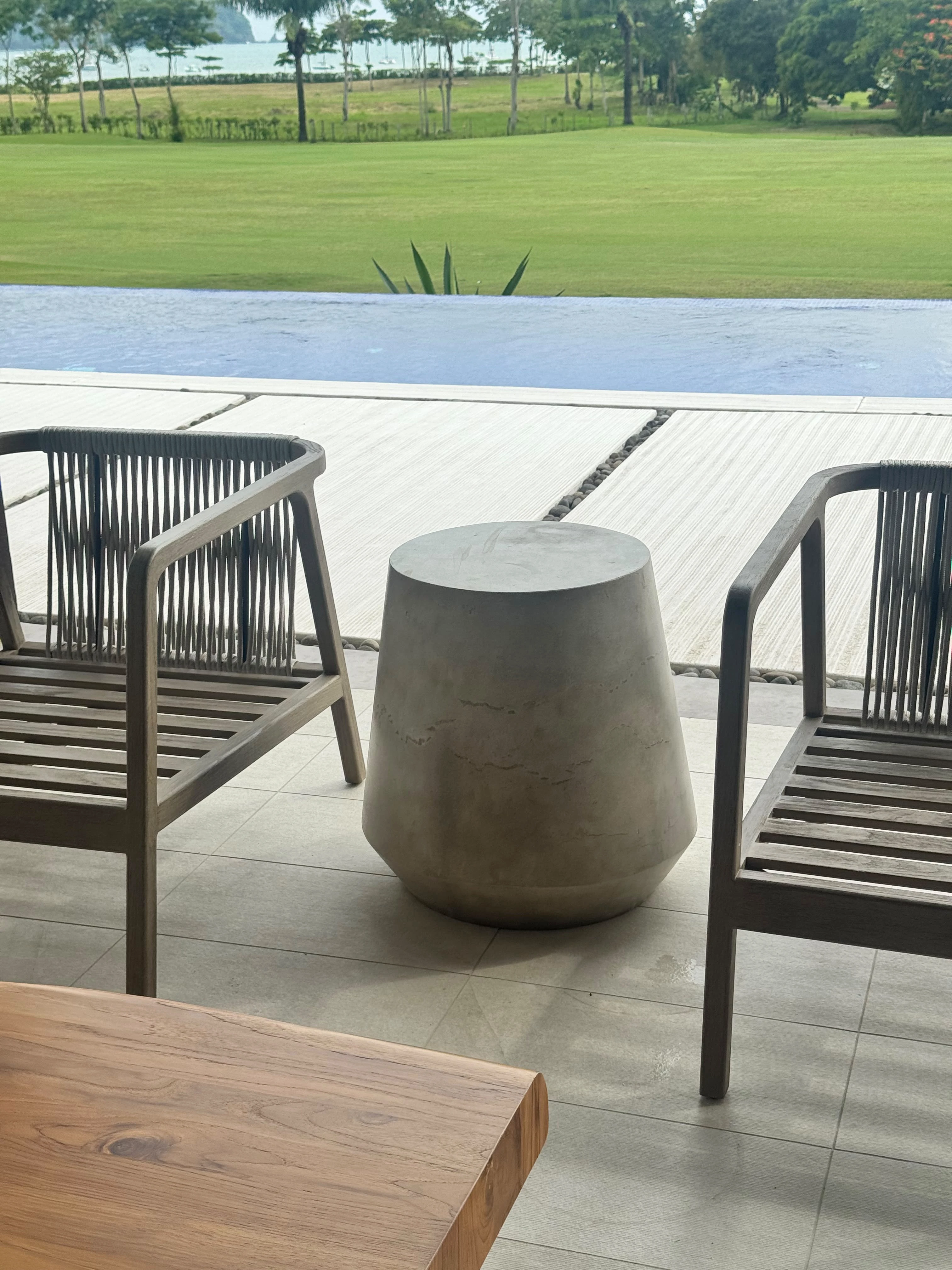 Banquito de concreto gris con forma cónica entre dos sillones de madera, en terraza junto a la piscina