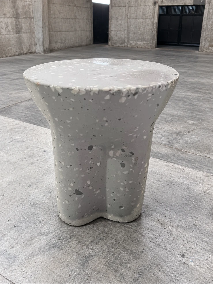 Banquito de terrazo blanco con agregados grises, tapa redonda y pie ensanchado con muescas, sobre piso de cemento pulido