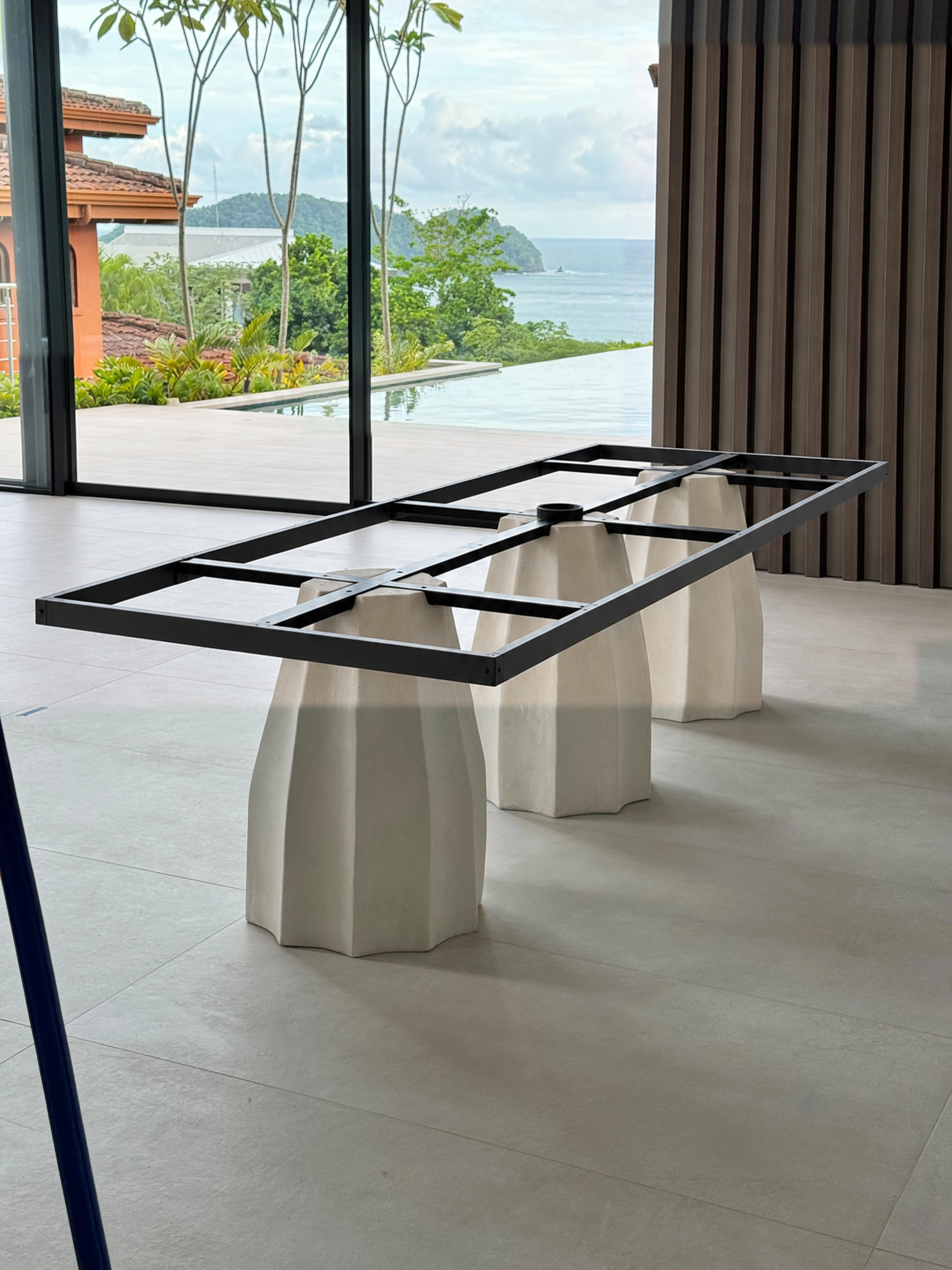 Base de mesa con tres pedestales de concreto blanco facetados y marco metálico negro, en sala con vista al mar