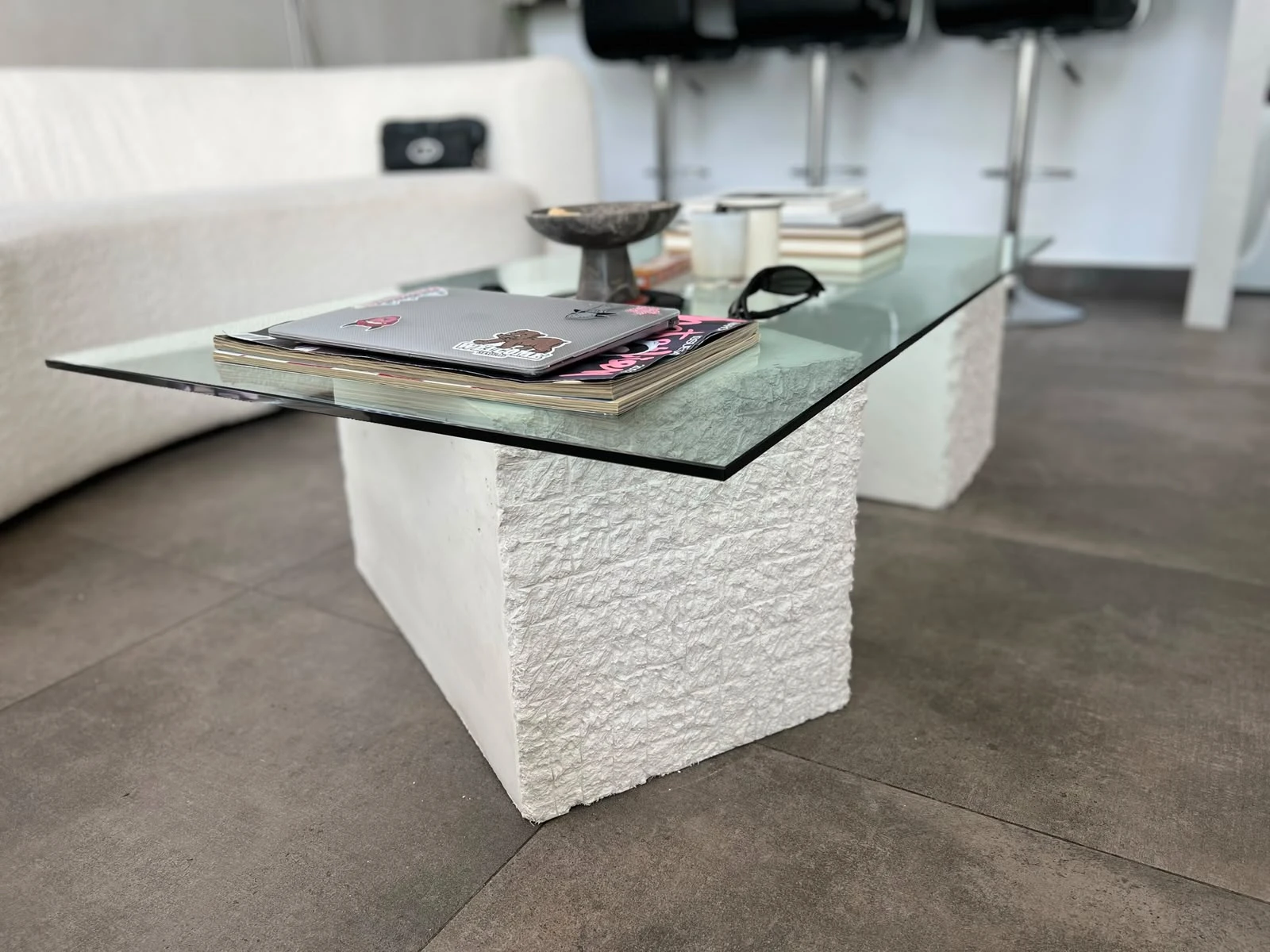 Mesa de centro con tapa de vidrio sobre bloques de concreto blanco de textura rugosa, frente a un sofá curvo