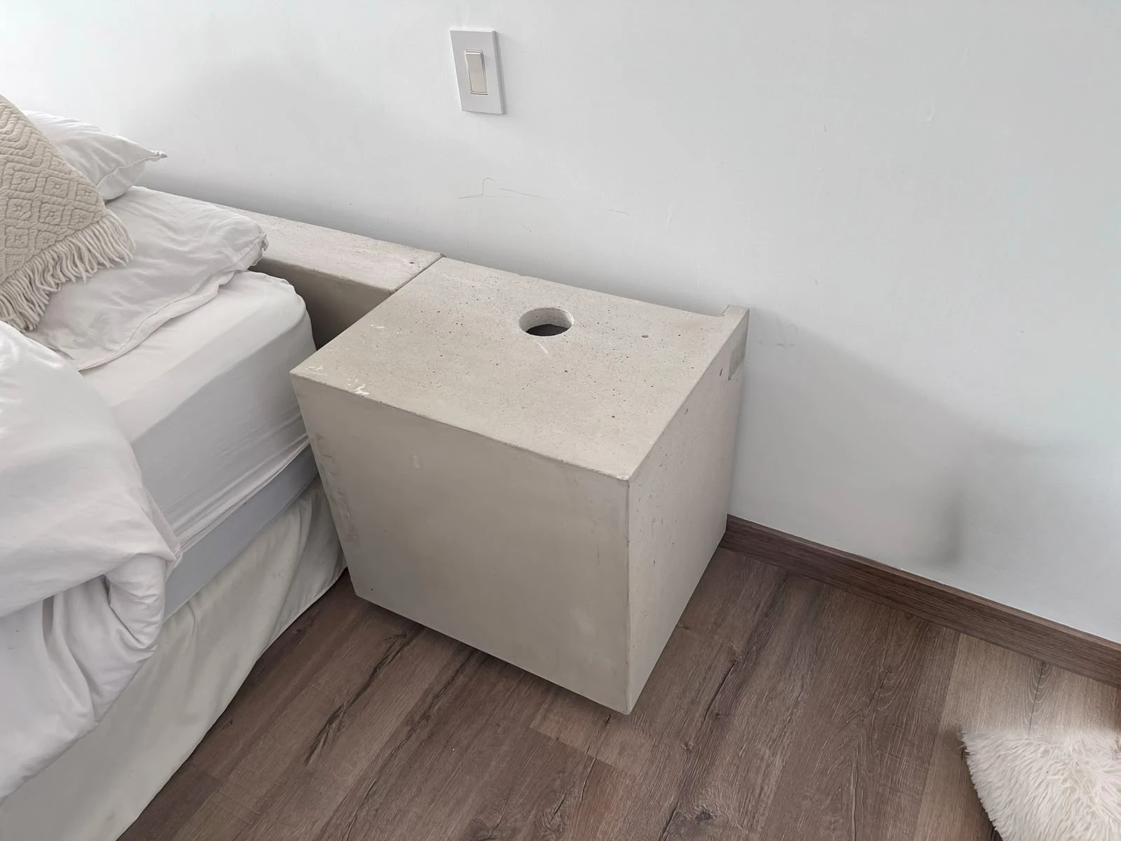 Mesa de noche cúbica de concreto claro con un orificio circular en la tapa, junto a una cama sobre piso de madera