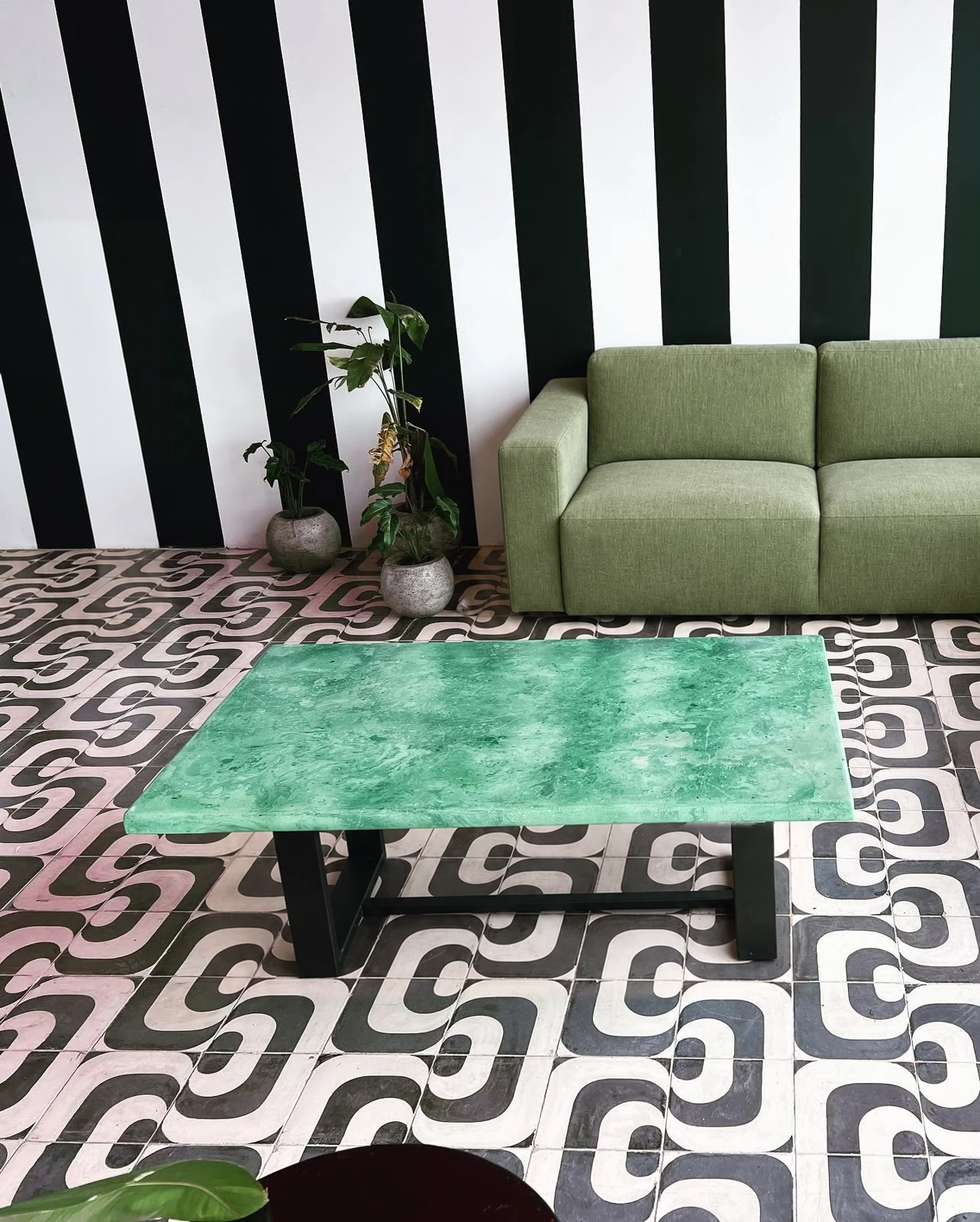 Mesa de centro con tapa de concreto verde jade y base de metal negro, sobre piso de mosaico en blanco y negro