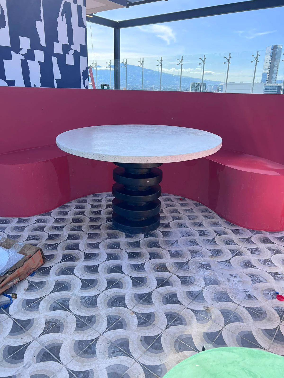 Mesa redonda de comedor con tapa de concreto blanco y base negra con anillos, entre bancas rosadas y piso de mosaico