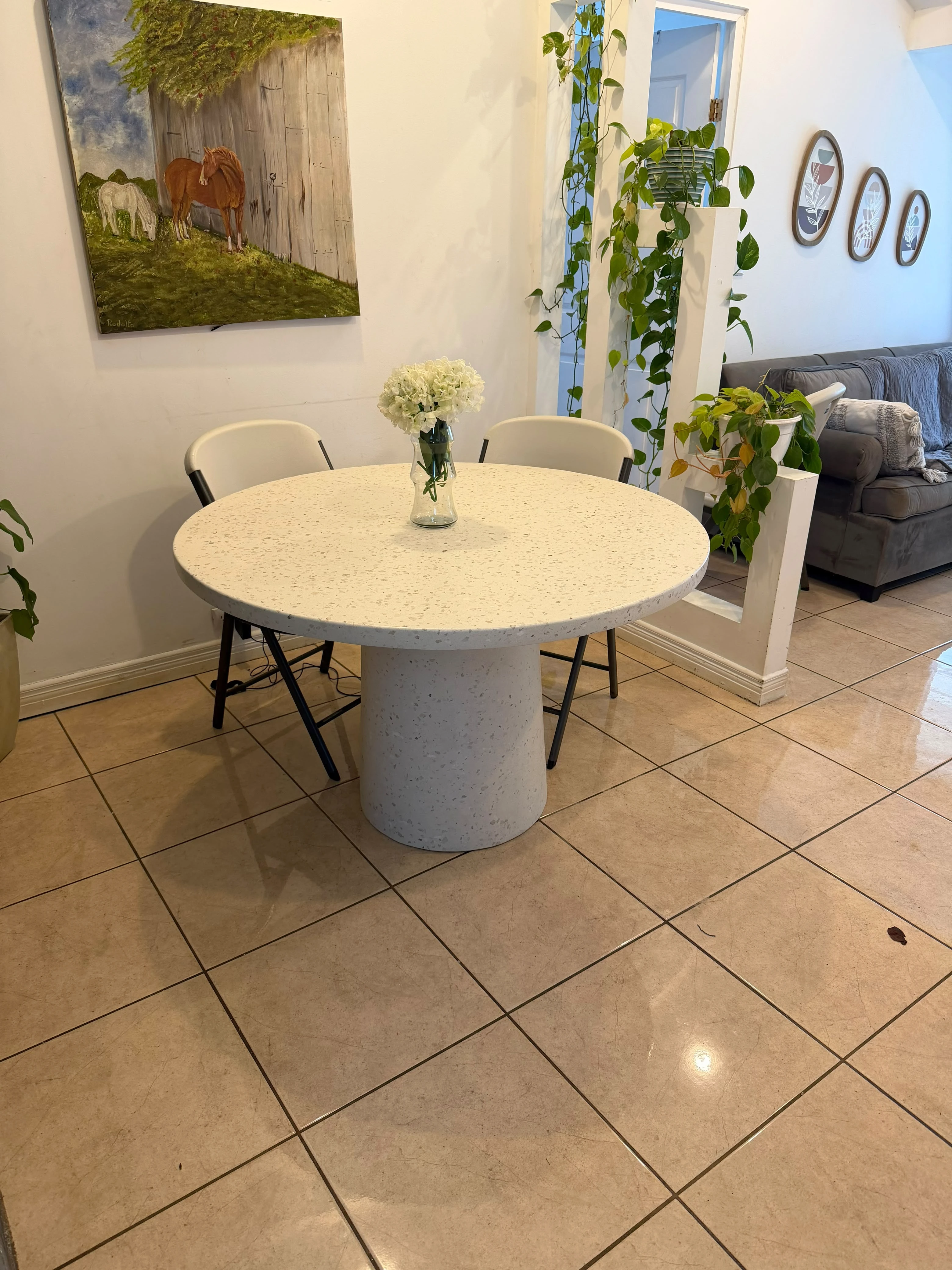 Mesa de comedor redonda de terrazo blanco con base cilíndrica, con florero, en comedor con piso de cerámica