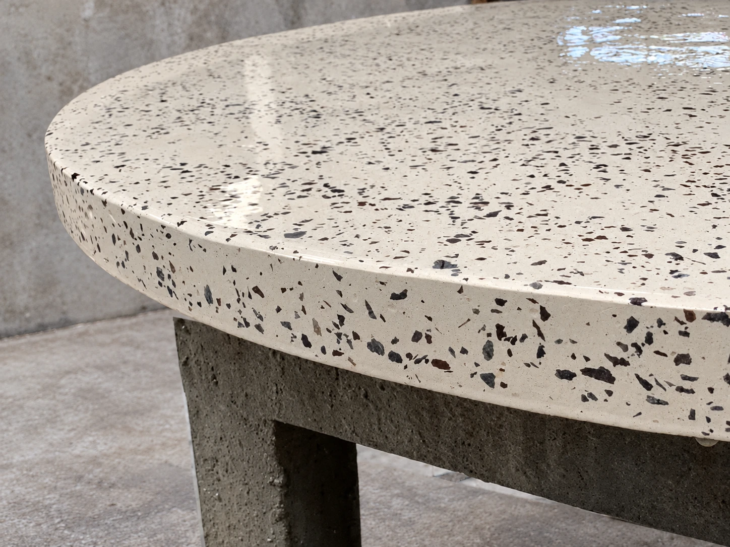 Detalle del borde de una mesa de terrazo blanco con agregados negros y acabado brillante, sobre pata de concreto gris