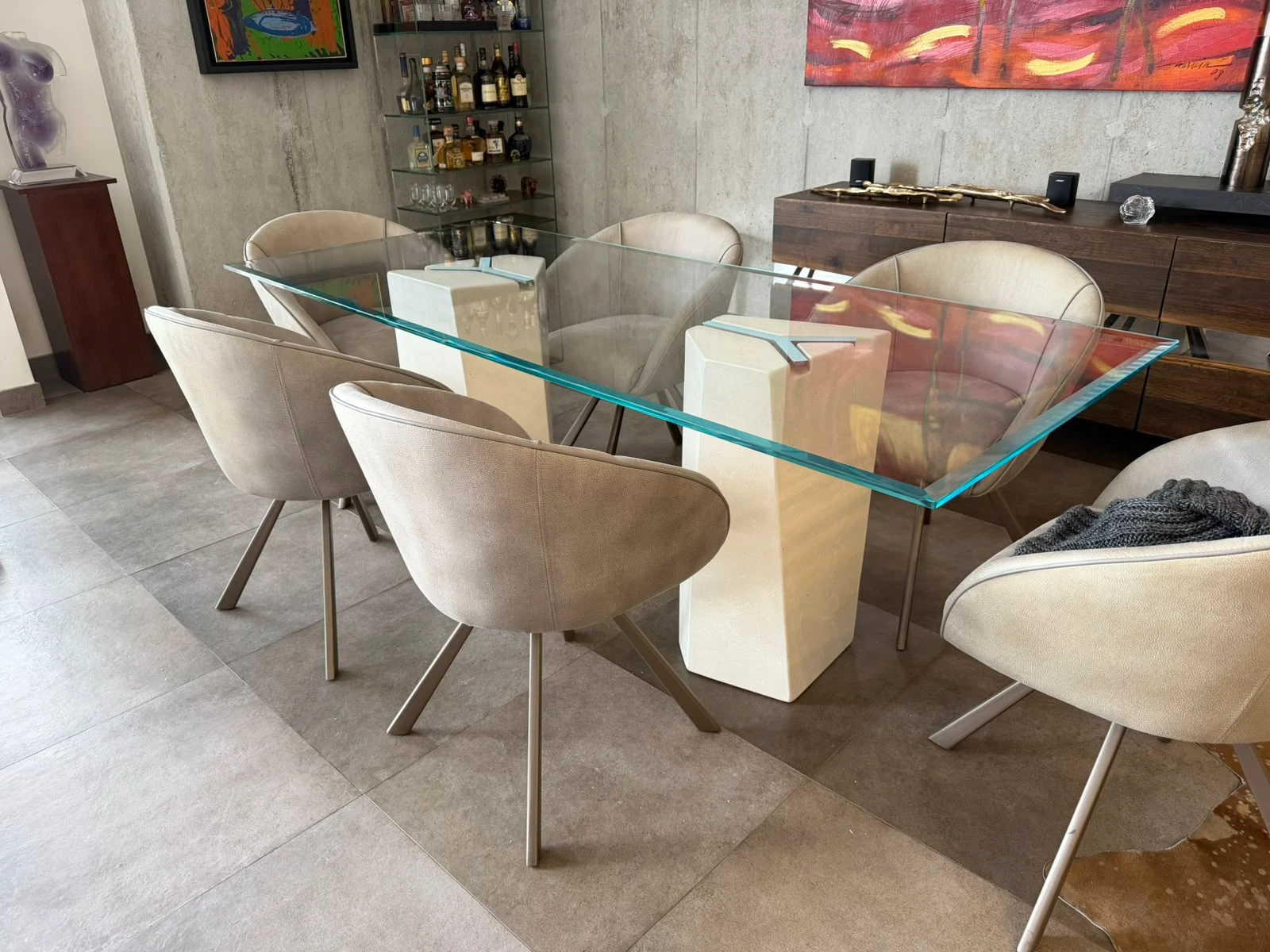 Mesa de comedor con tapa de vidrio sobre dos pedestales de concreto crema, rodeada de sillas tapizadas en sala amplia