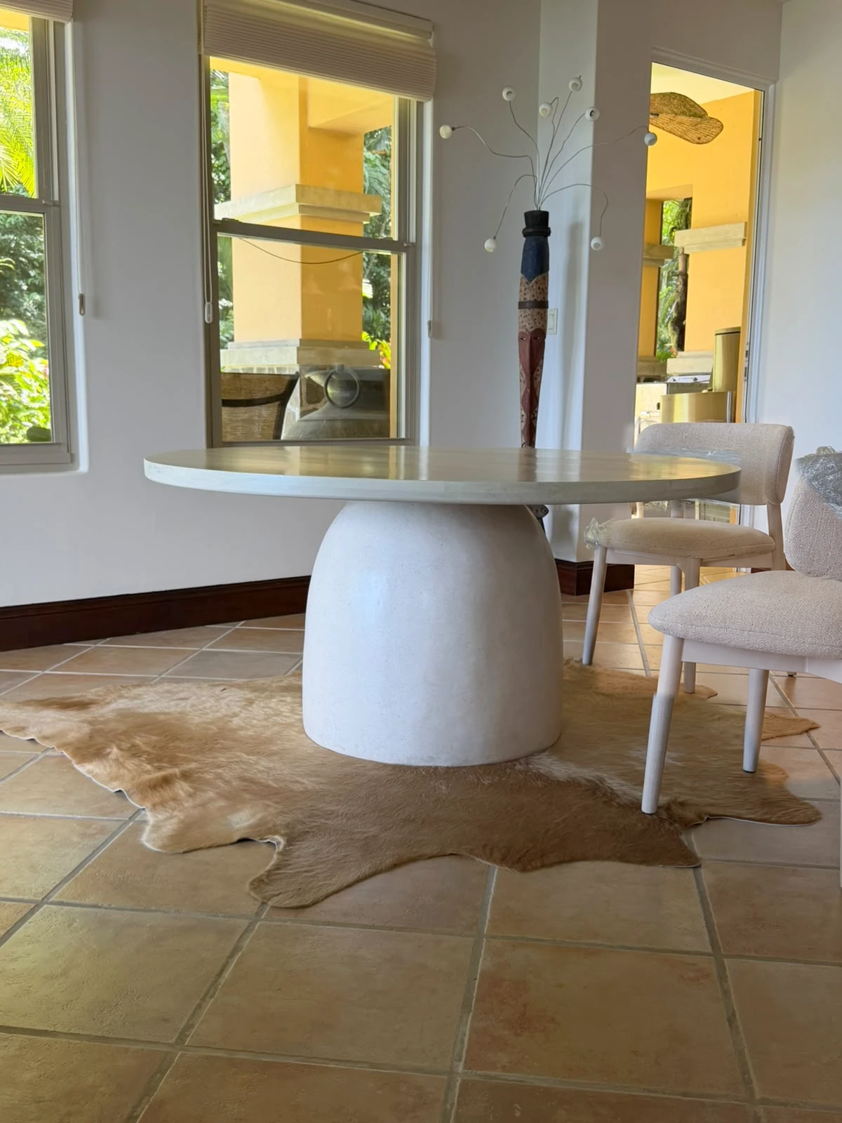 Mesa de comedor redonda con tapa de concreto blanco y pie en forma de campana, sobre alfombra de cuero y piso de barro
