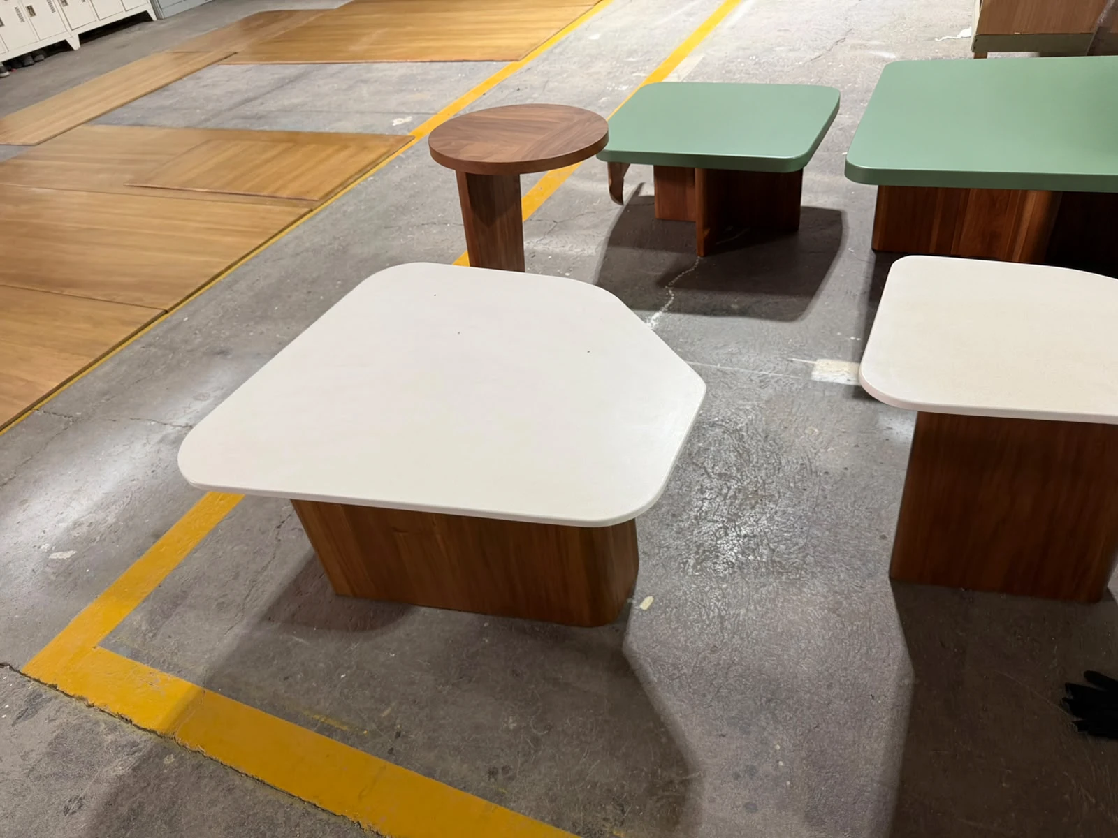 Mesas bajas con tapas blancas de formas irregulares y bases de madera, junto a mesitas verdes, sobre piso del taller