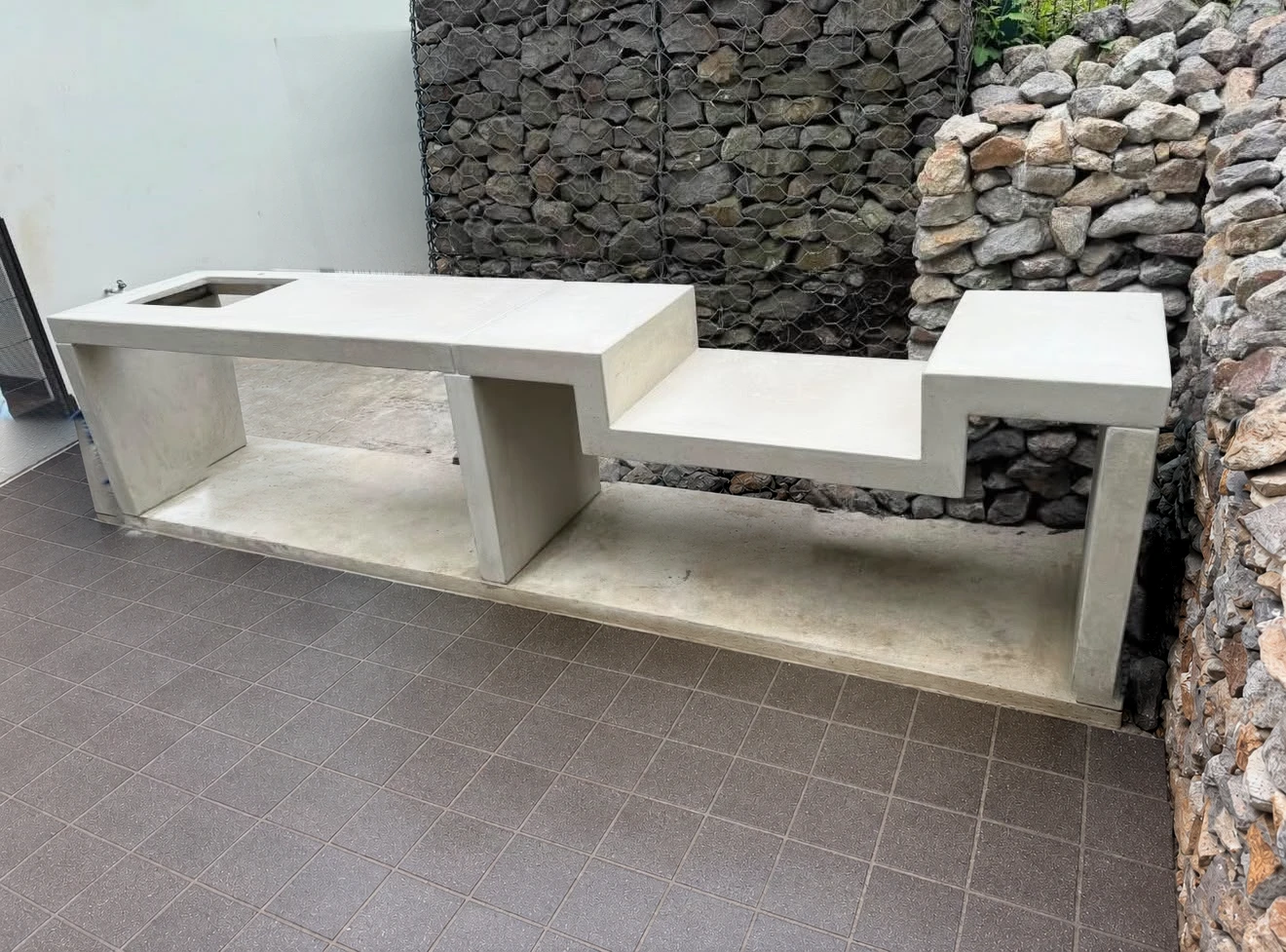 Mueble de concreto claro en dos niveles con pila rectangular en un extremo, junto a muro de piedra en patio exterior