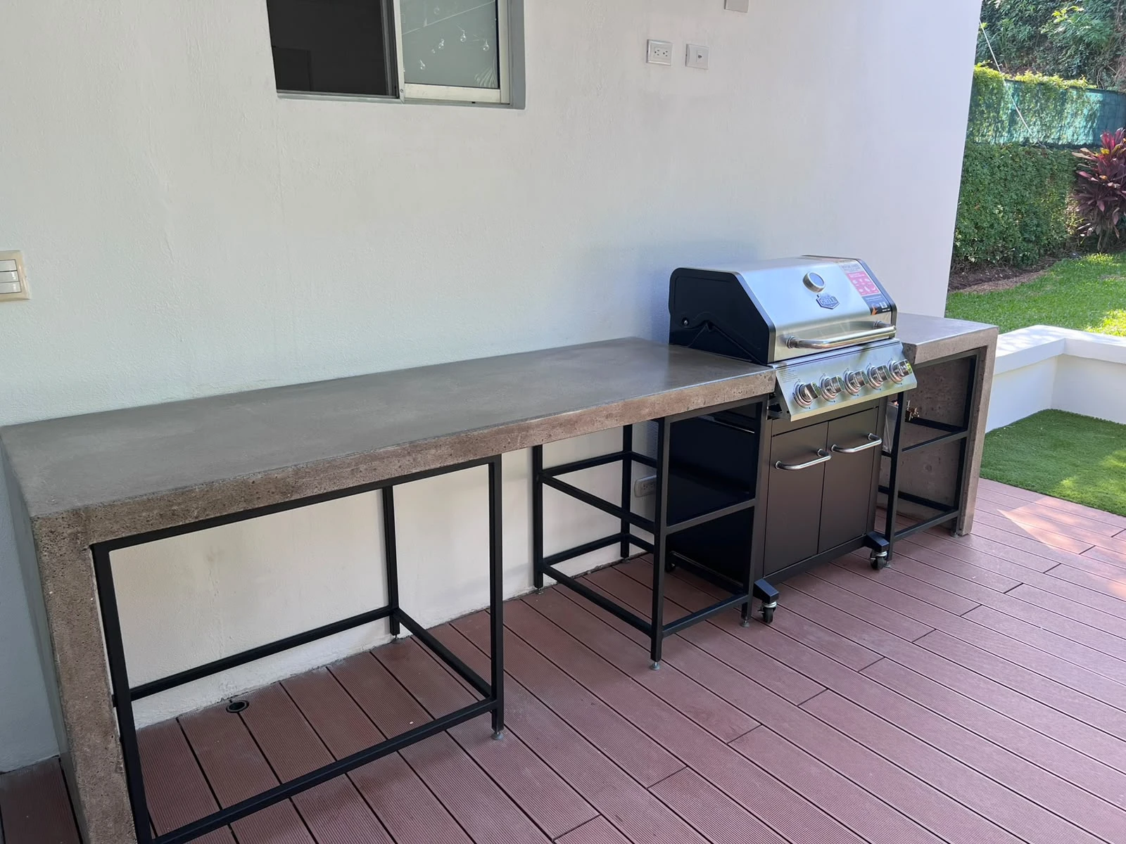 Sobre largo de concreto gris sobre estructura metálica negra, con parrilla de gas integrada, en terraza con deck rosado