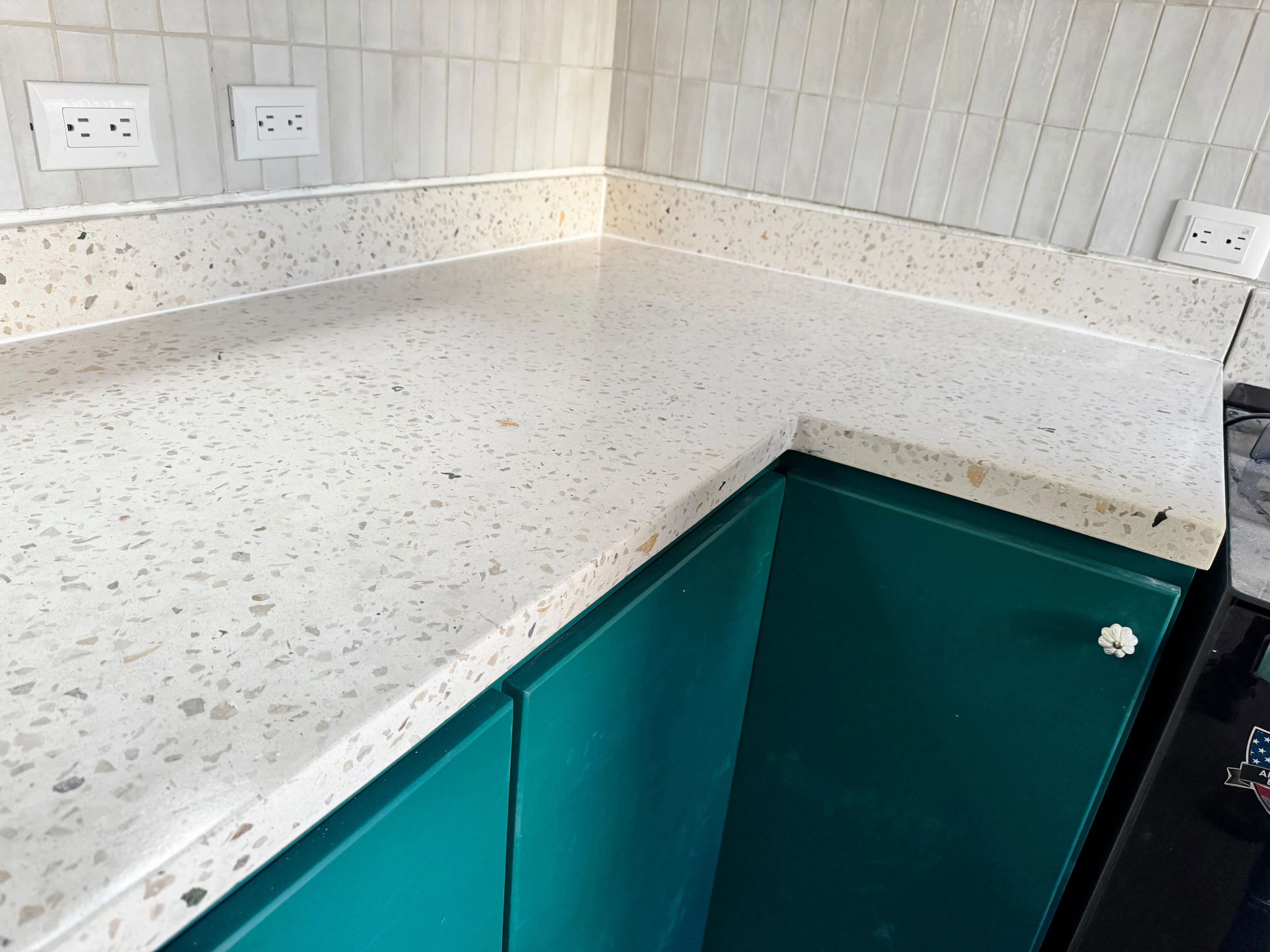 Sobre de cocina en forma de L de terrazo claro con agregados oscuros y zócalo del mismo material, sobre mueble verde azulado