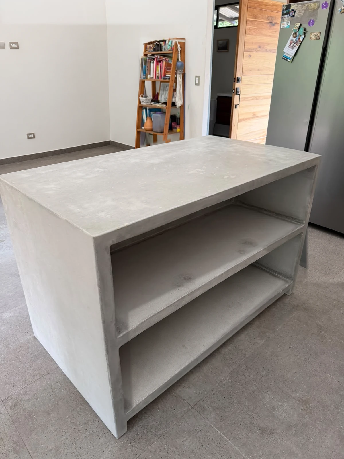 Mueble tipo isla de concreto gris claro con sobre liso y dos repisas abiertas, en un espacio con piso de cemento