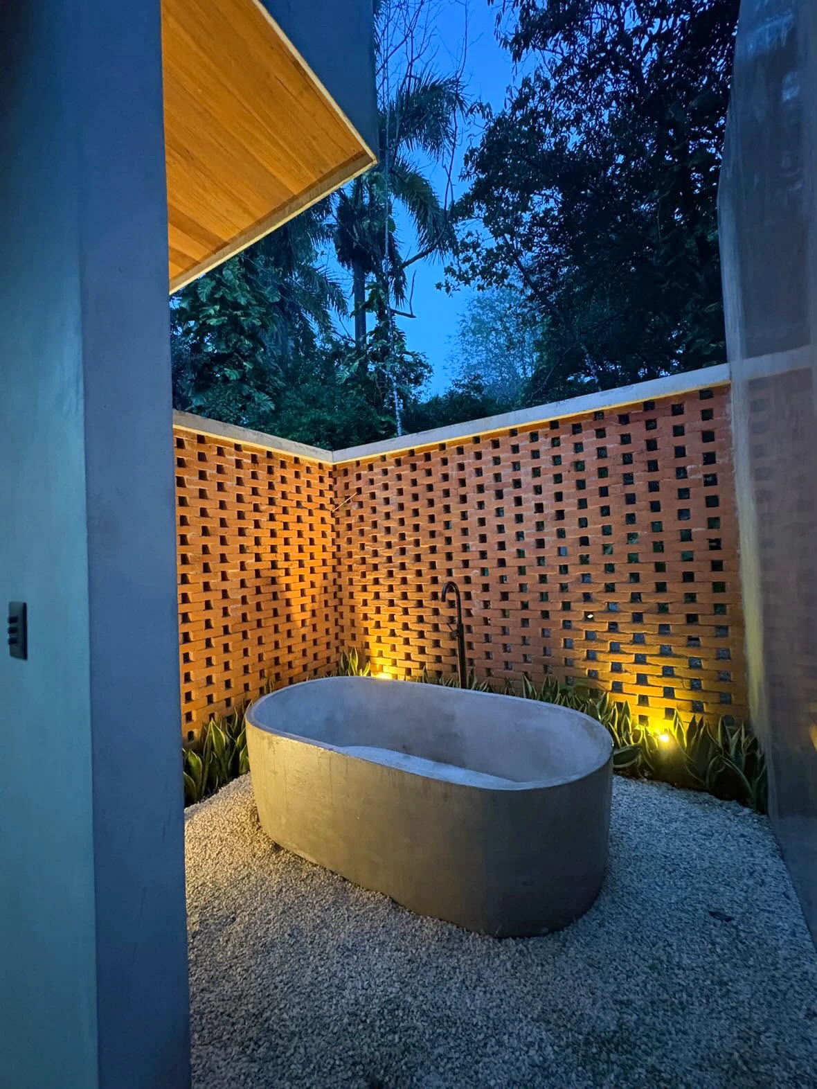 Tina ovalada de concreto gris exenta sobre grava, en patio exterior con muro de ladrillo calado e iluminación cálida al atardecer