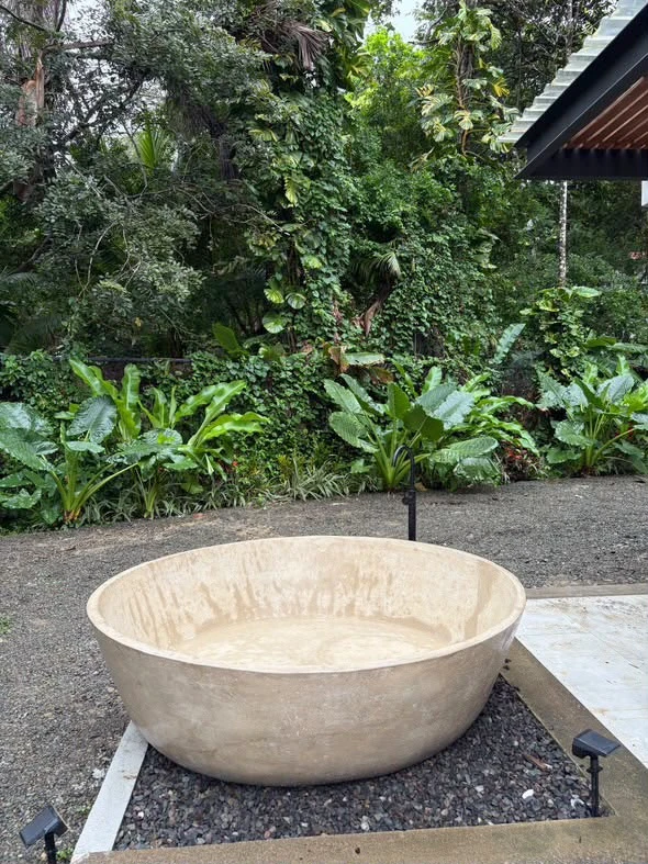 Tina redonda de concreto color travertino con borde alto, instalada sobre grava en jardín rodeado de vegetación tropical