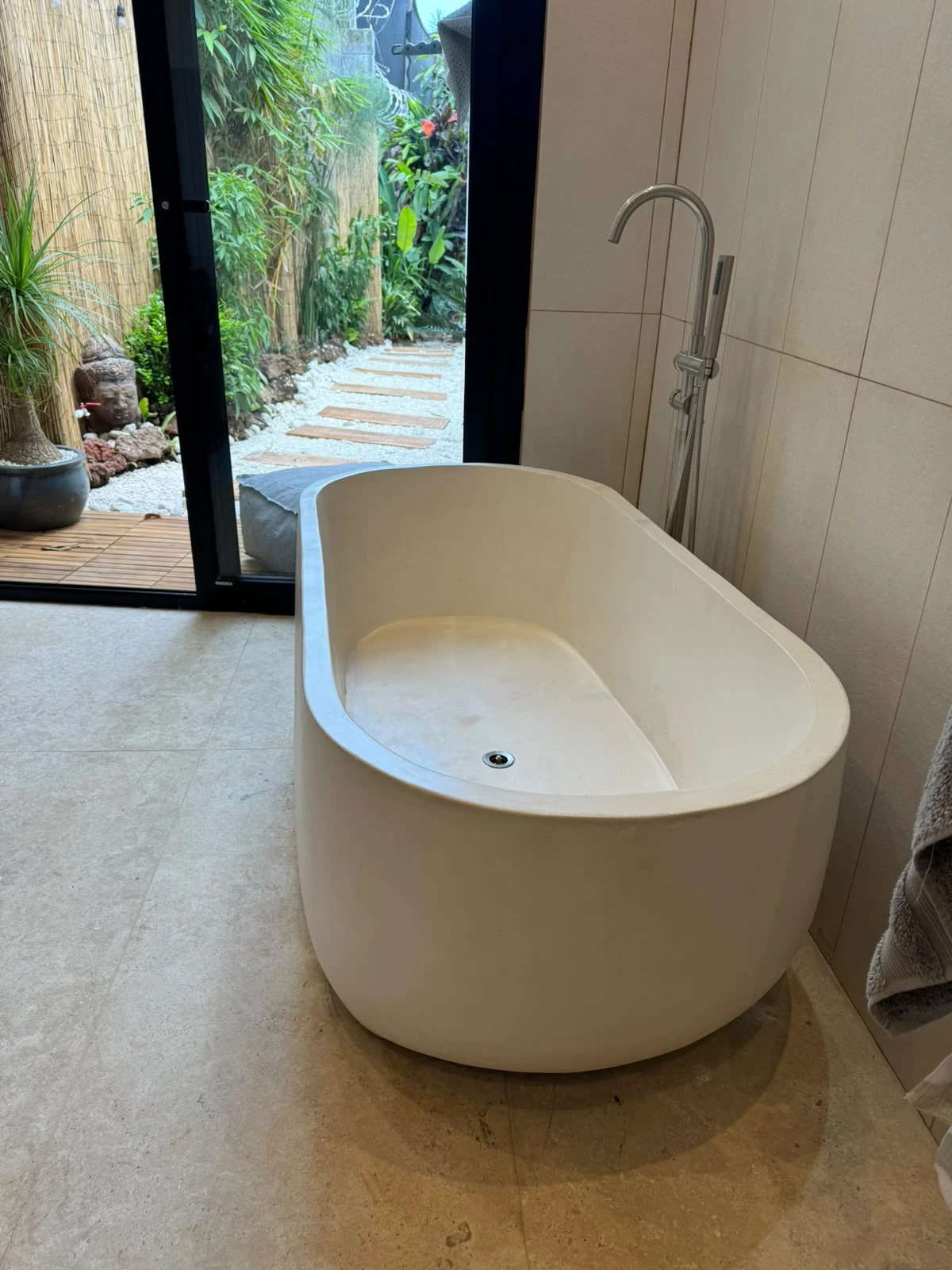Tina ovalada exenta de concreto blanco con borde redondeado, en baño con puerta de vidrio hacia un jardín