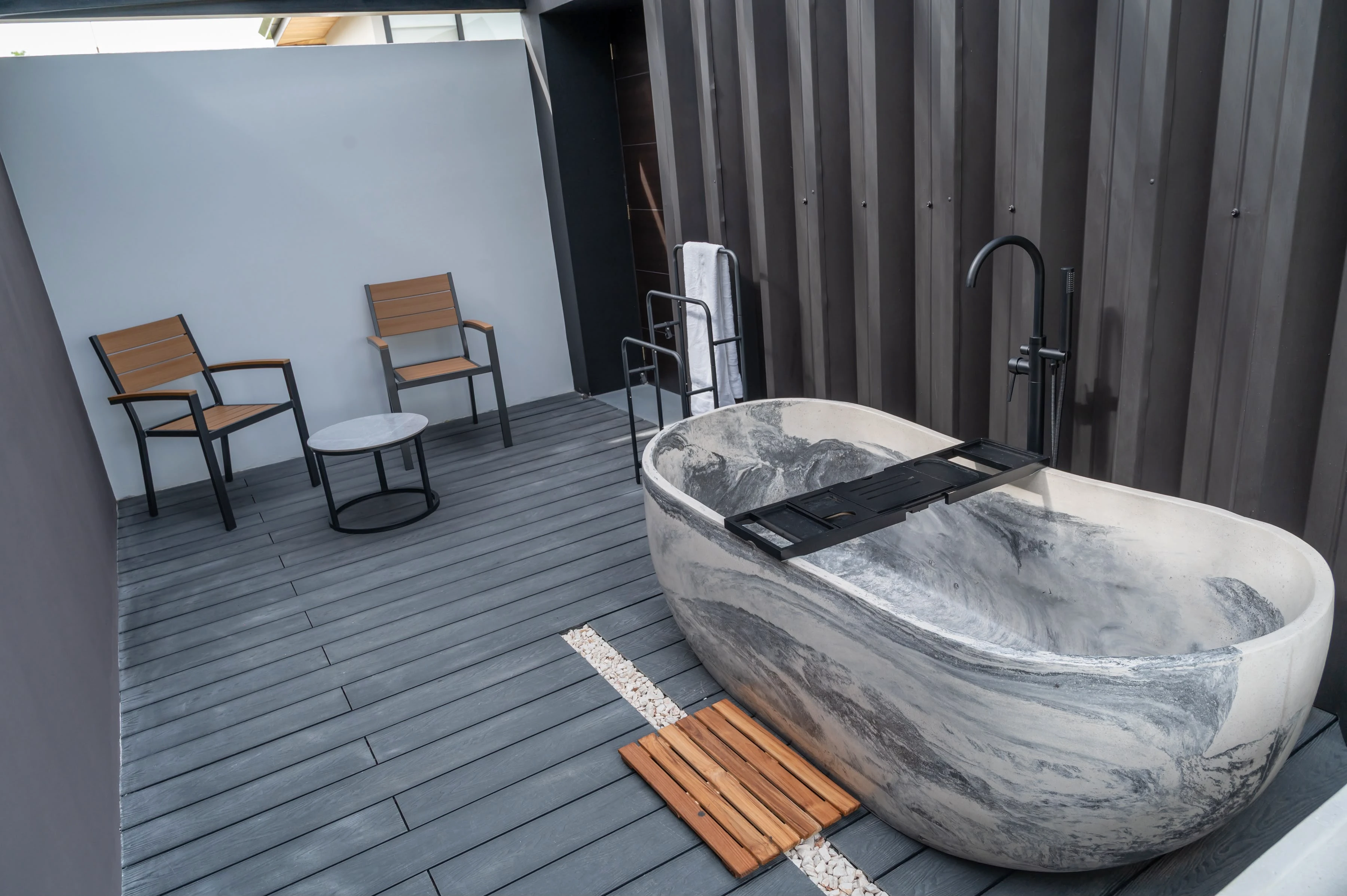 Tina ovalada de concreto marmoleado gris y blanco con bandeja negra, sobre terraza de deck con sillas de madera