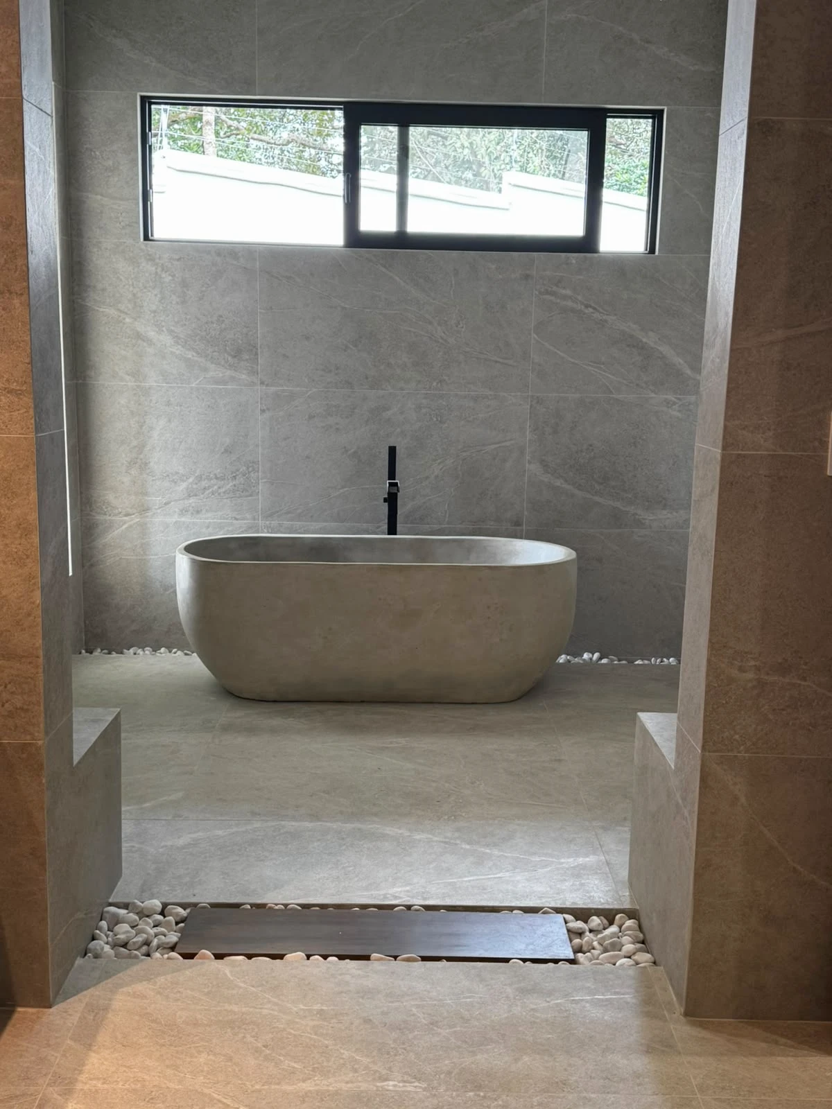 Tina ovalada exenta de concreto gris con grifería negra de piso, en baño con paredes de porcelanato gris y piedras de río